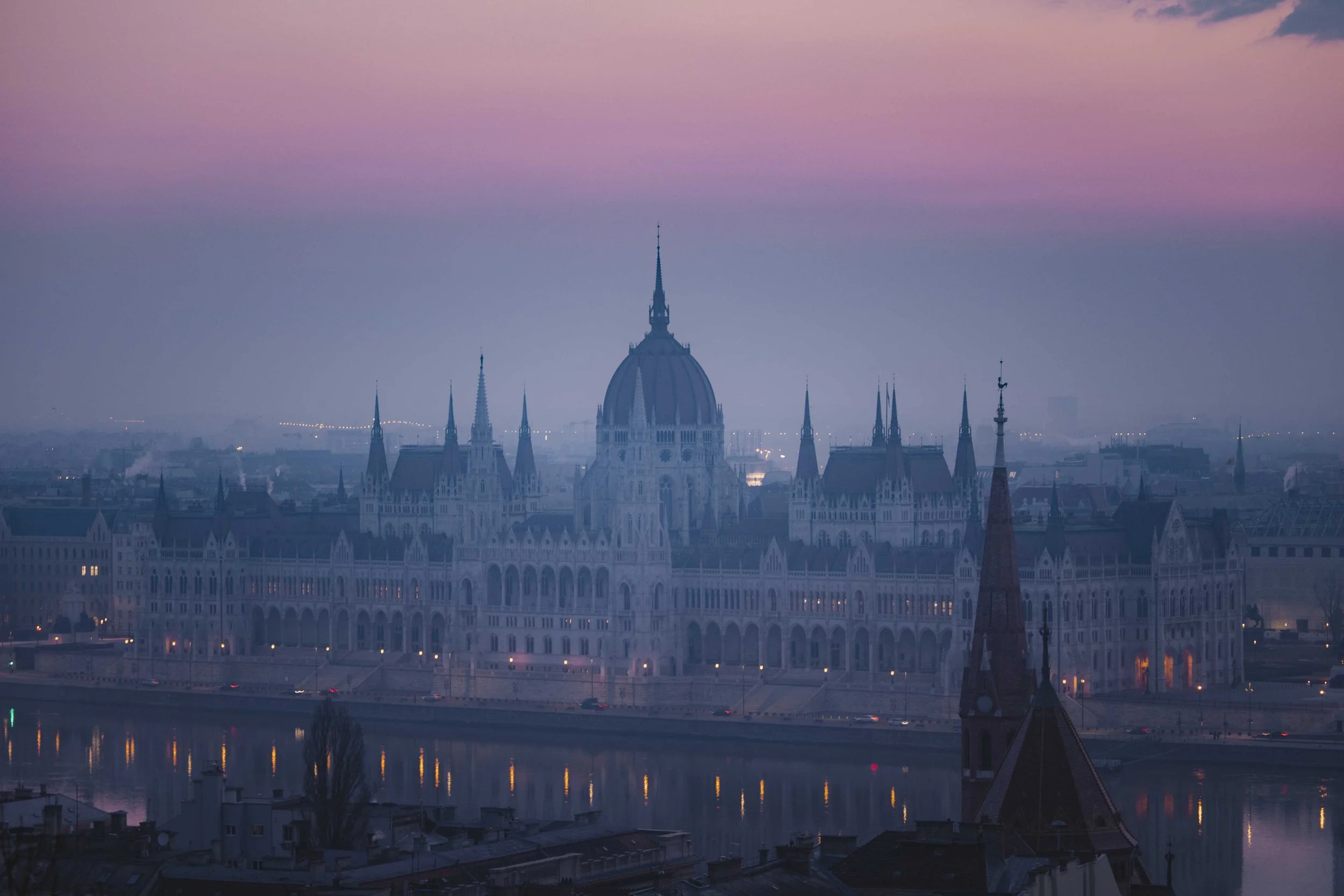 The Parliament building in Budapest, Hungary, seen during dusk with a pastel sky and the Danube River in the foreground.