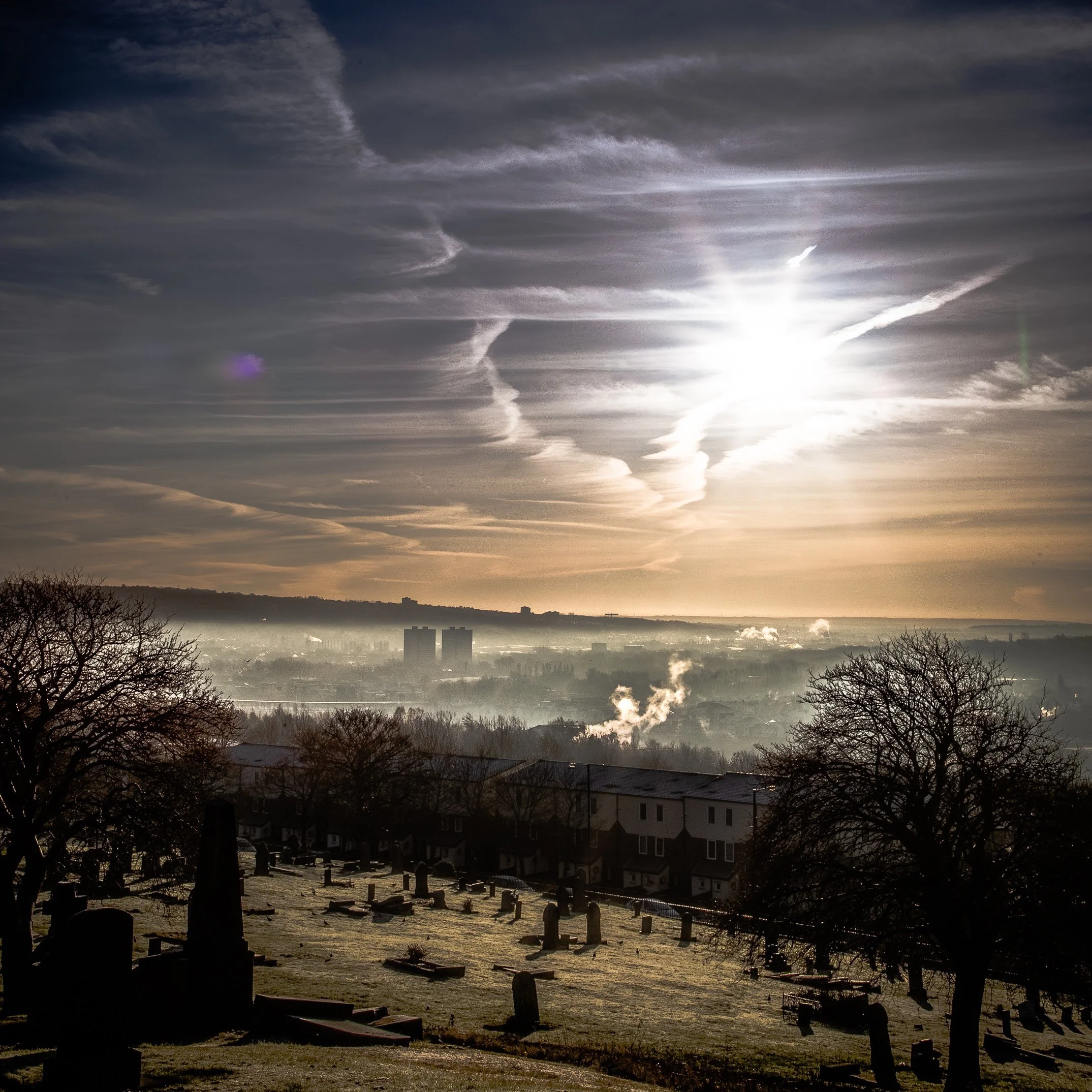 A sunrise over a cemetery with tombstones and leafless trees, with a cityscape and hills in the background and a partly cloudy sky.