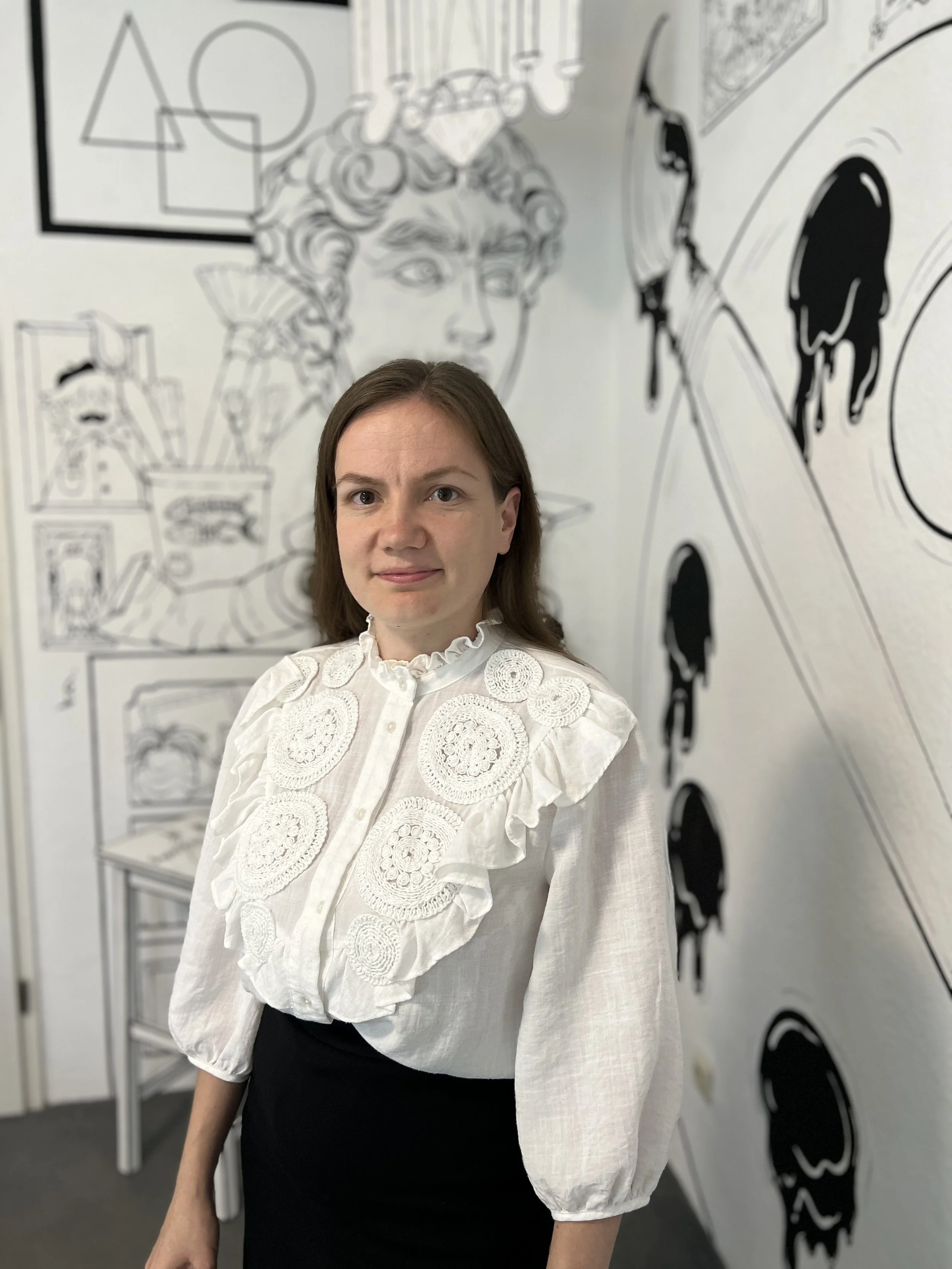 Eine Frau mit braunen Haaren in einem weißen, gestrickten Bluse mit Rüschen steht vor einer Wand mit Schwarz-Weiß-Illustrationen, darunter eine große Darstellung einer antiken Statue einer Medusa-ähnlichen Figur, und anderen Kunstwerken.