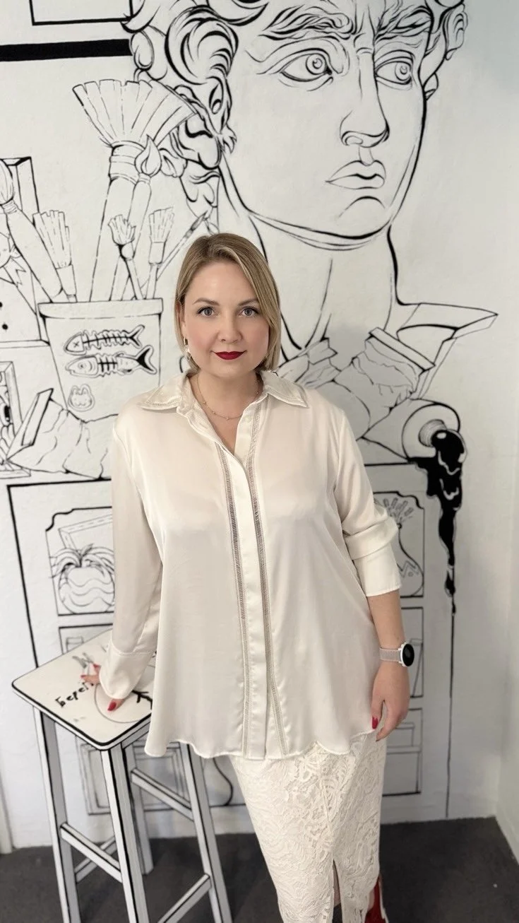 Eine Frau steht vor einer schwarz-weißen Wandzeichnung, die eine große Kultstatue mit gewelltem Haar und ausdrucksstarker Mimik zeigt. Im Hintergrund sind Pinsel und Zeichenutensilien in einem Becher sichtbar.