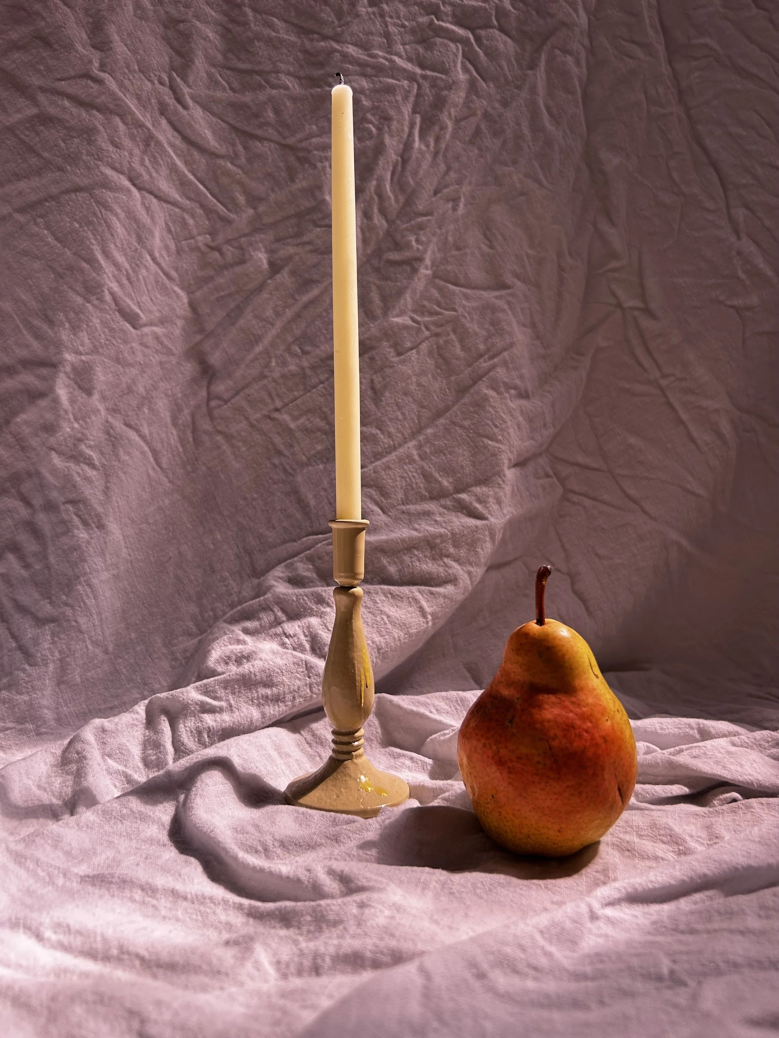 A still life composition with a beige candlestick holder and a pear placed on a wrinkled, light pink fabric background.