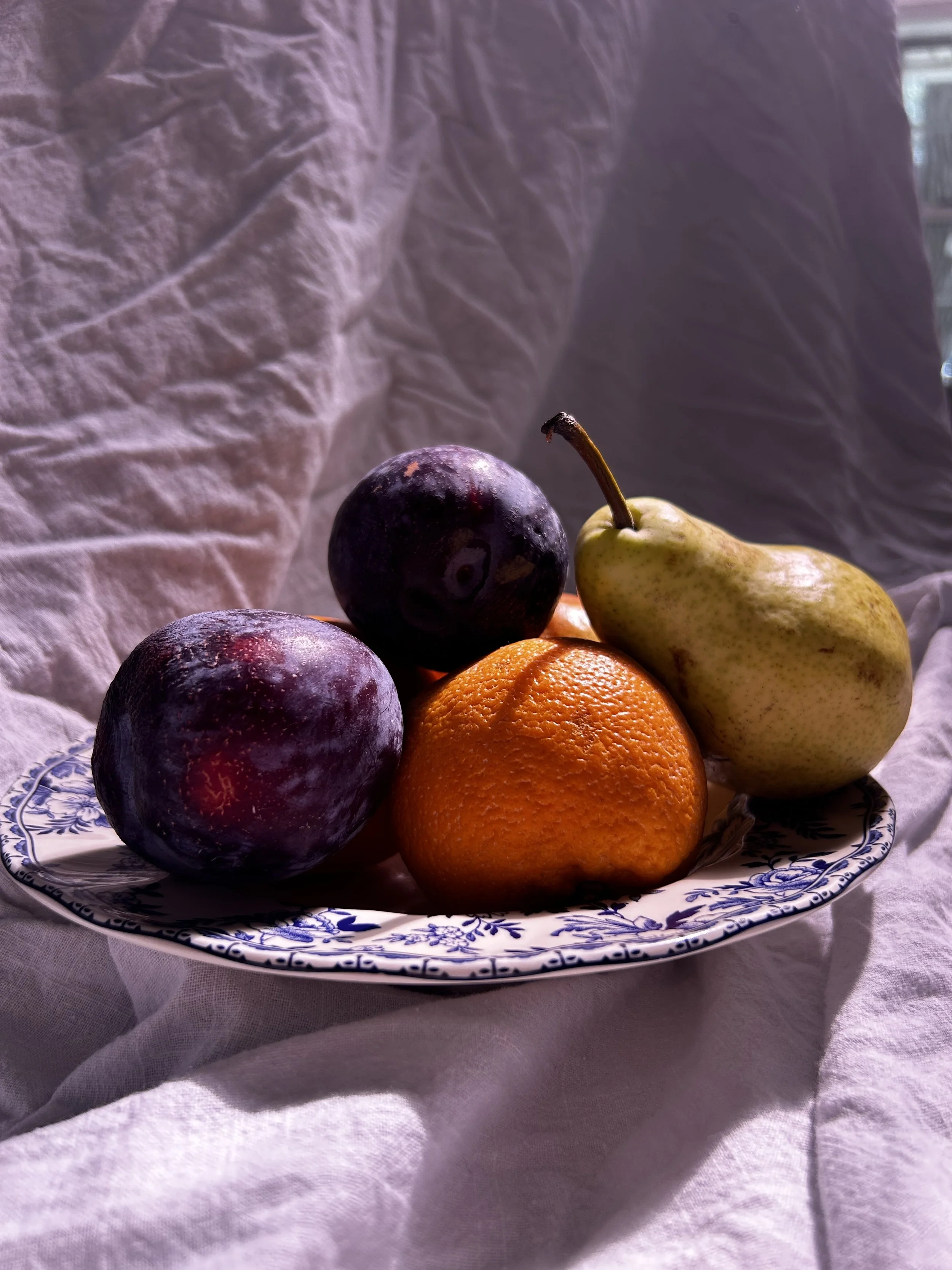A decorative plate with purple plums, a pear, and an orange, placed on a white cloth with soft lighting.