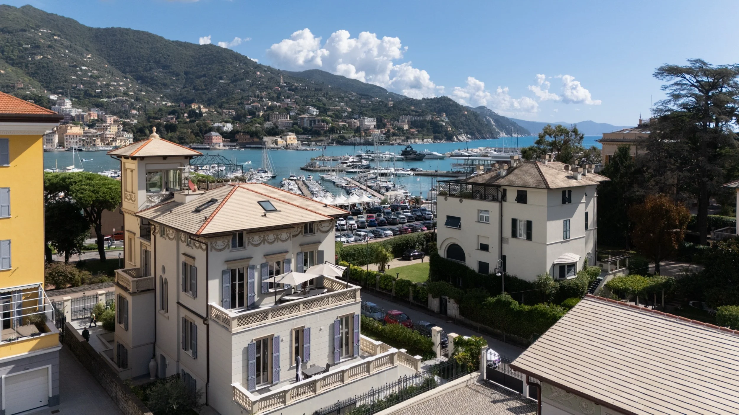 A scenic view of a marina with numerous boats and yachts docked on the water, surrounded by colorful residential buildings, lush green trees, and mountains in the background under a partly cloudy sky.