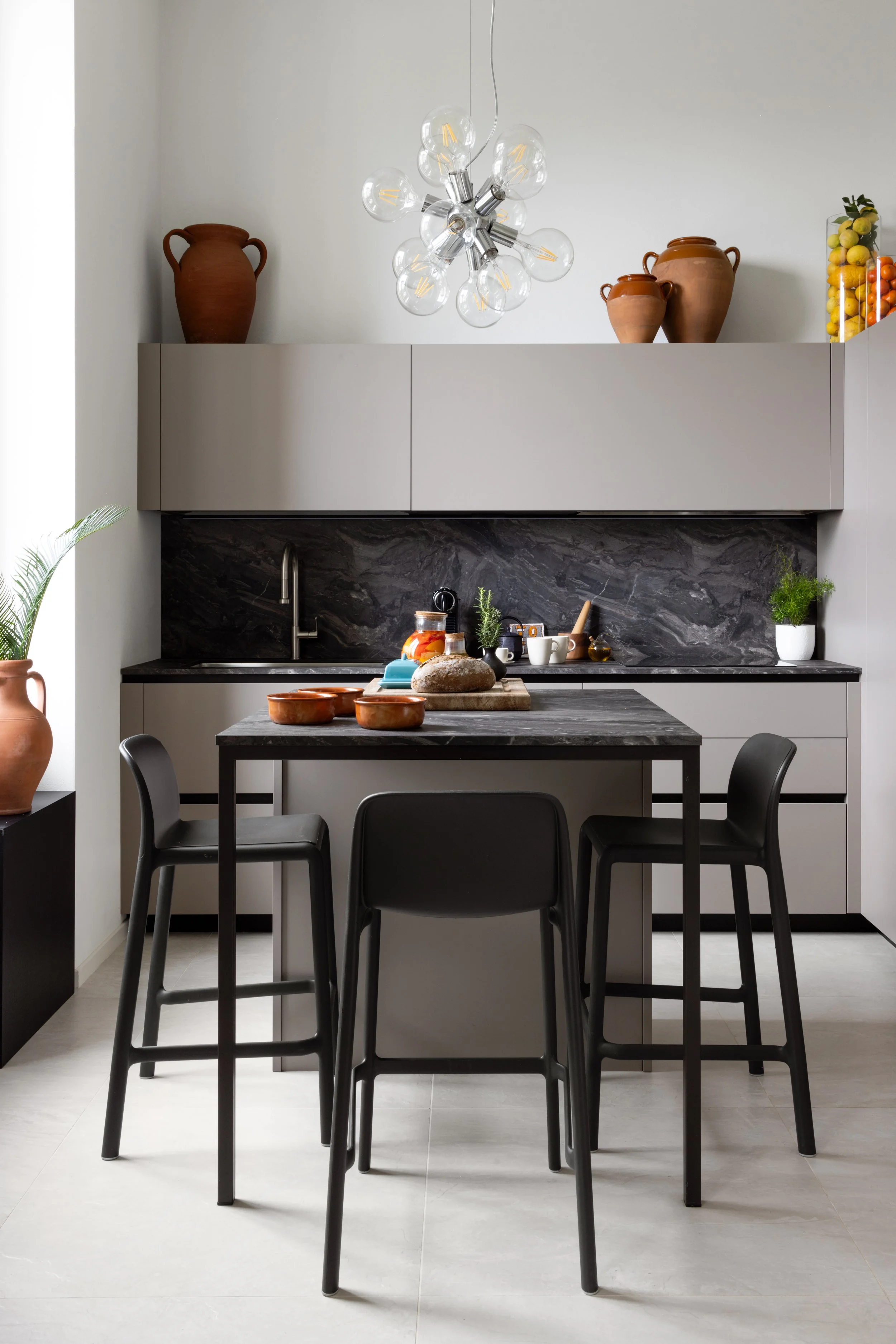 Modern kitchen with black table, four black chairs, gray cabinets, black marble backsplash, terracotta jars, plants, and a black and gold pendant light fixture.