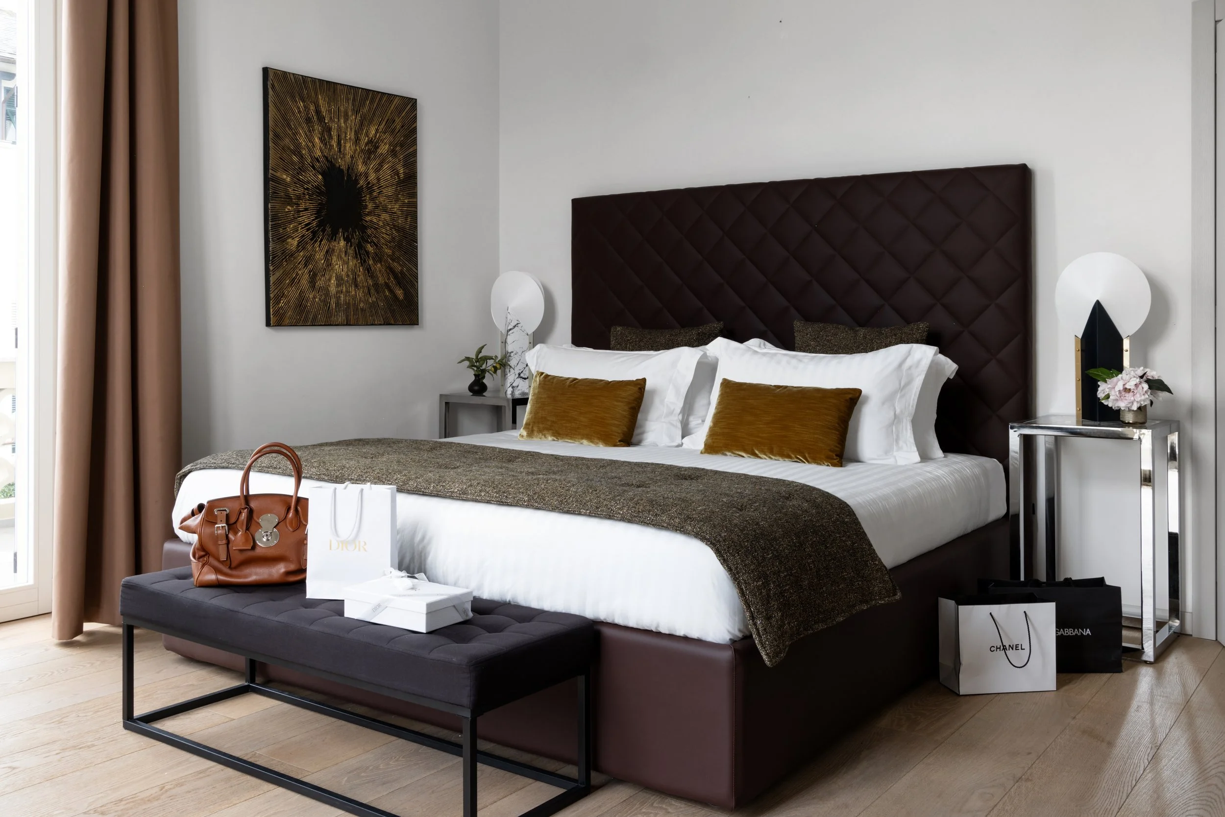 A neatly made bedroom with a bed featuring a large dark purple, diamond-tufted headboard, decorative pillows, and a brown blanket. Beside the bed are side tables with modern lamps, a small plant, and flowers. A large abstract artwork hangs on the whi