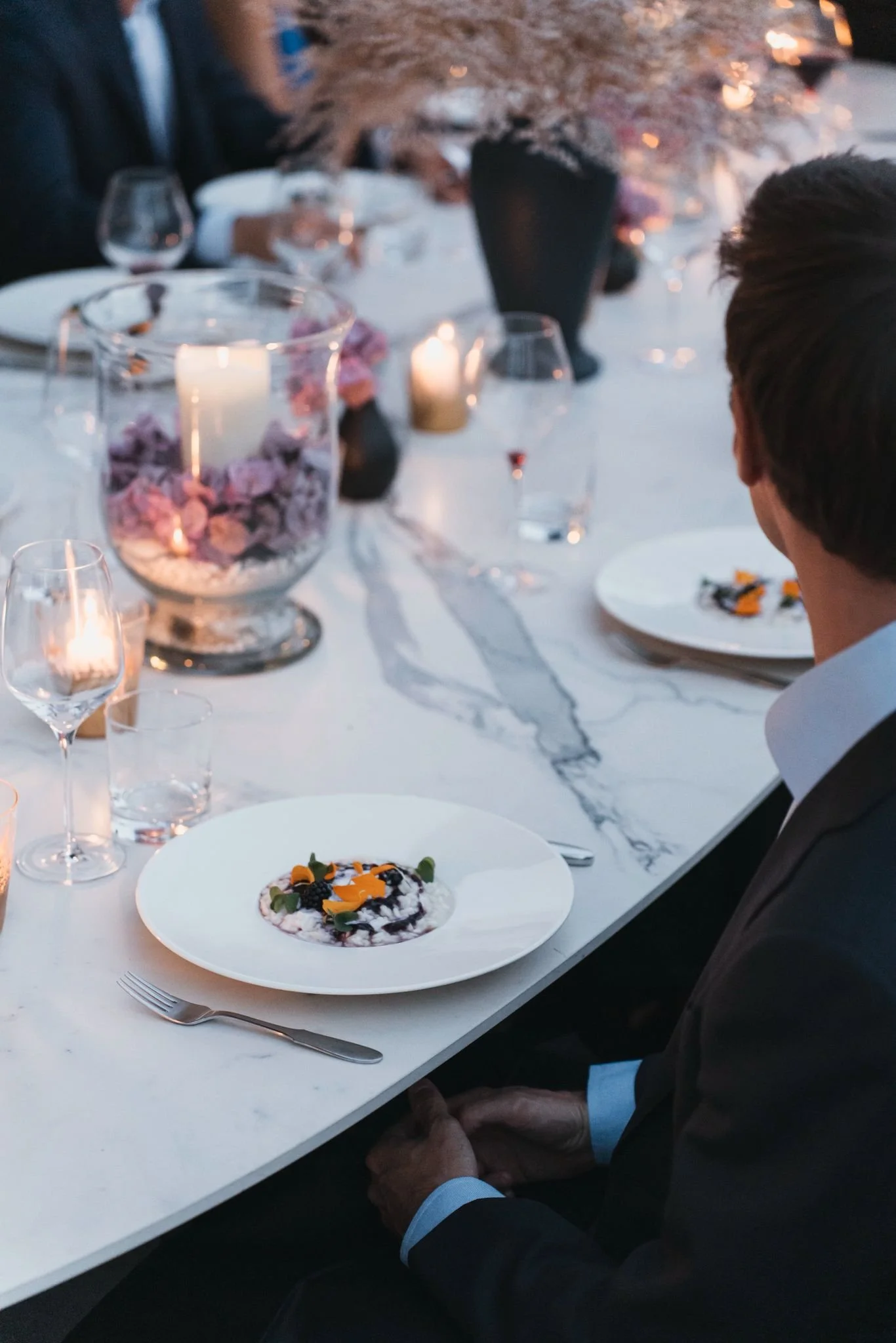 A dinner party scene with a marble table, glasses, candles, and a dessert with berries and edible flowers. A person in a dark suit is seated at the table, with only part of their face and upper body visible.
