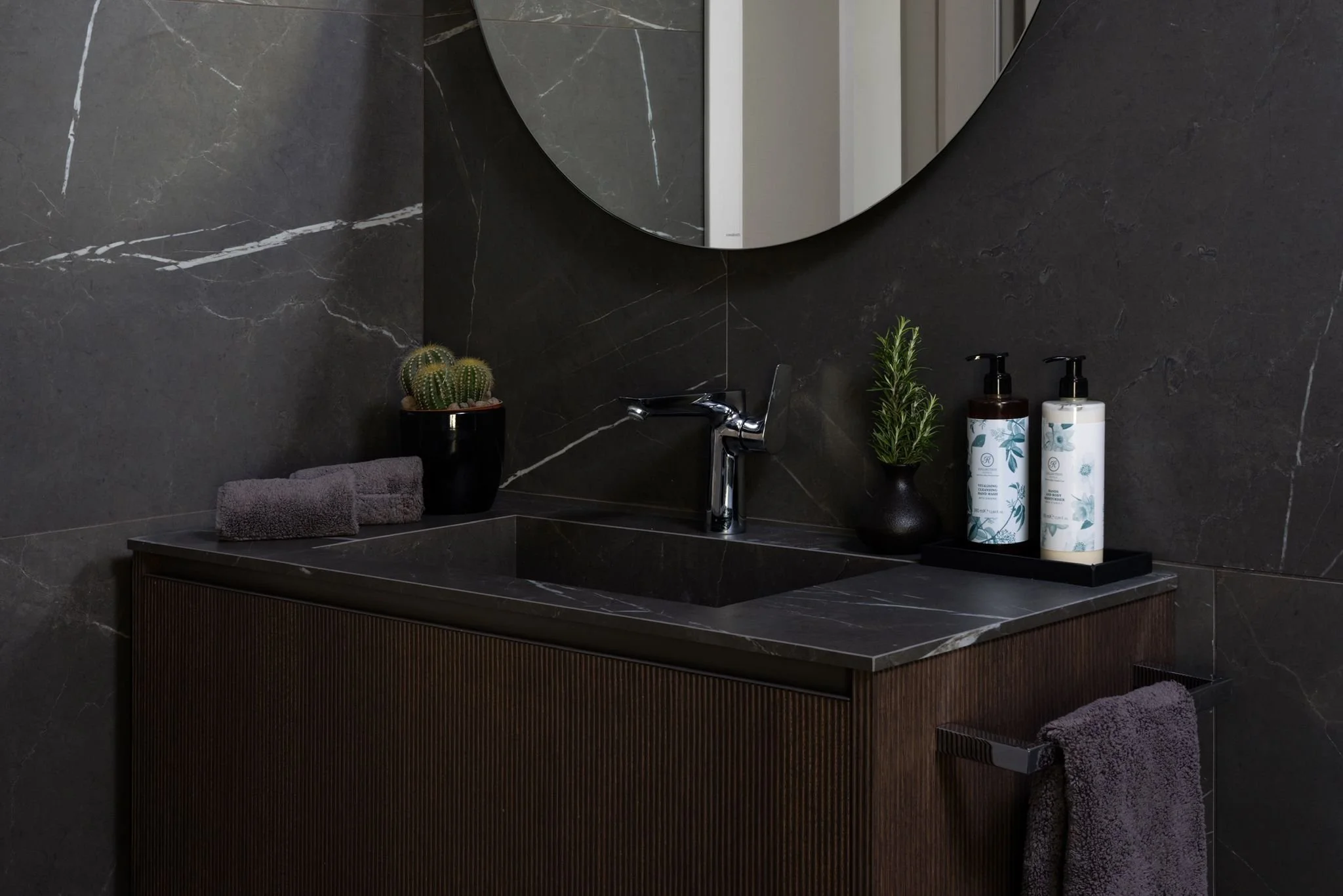A bathroom sink area with dark marble surfaces, a round mirror, a small potted cactus, a black tray with a rolled towel, and two bottles of hand soap.