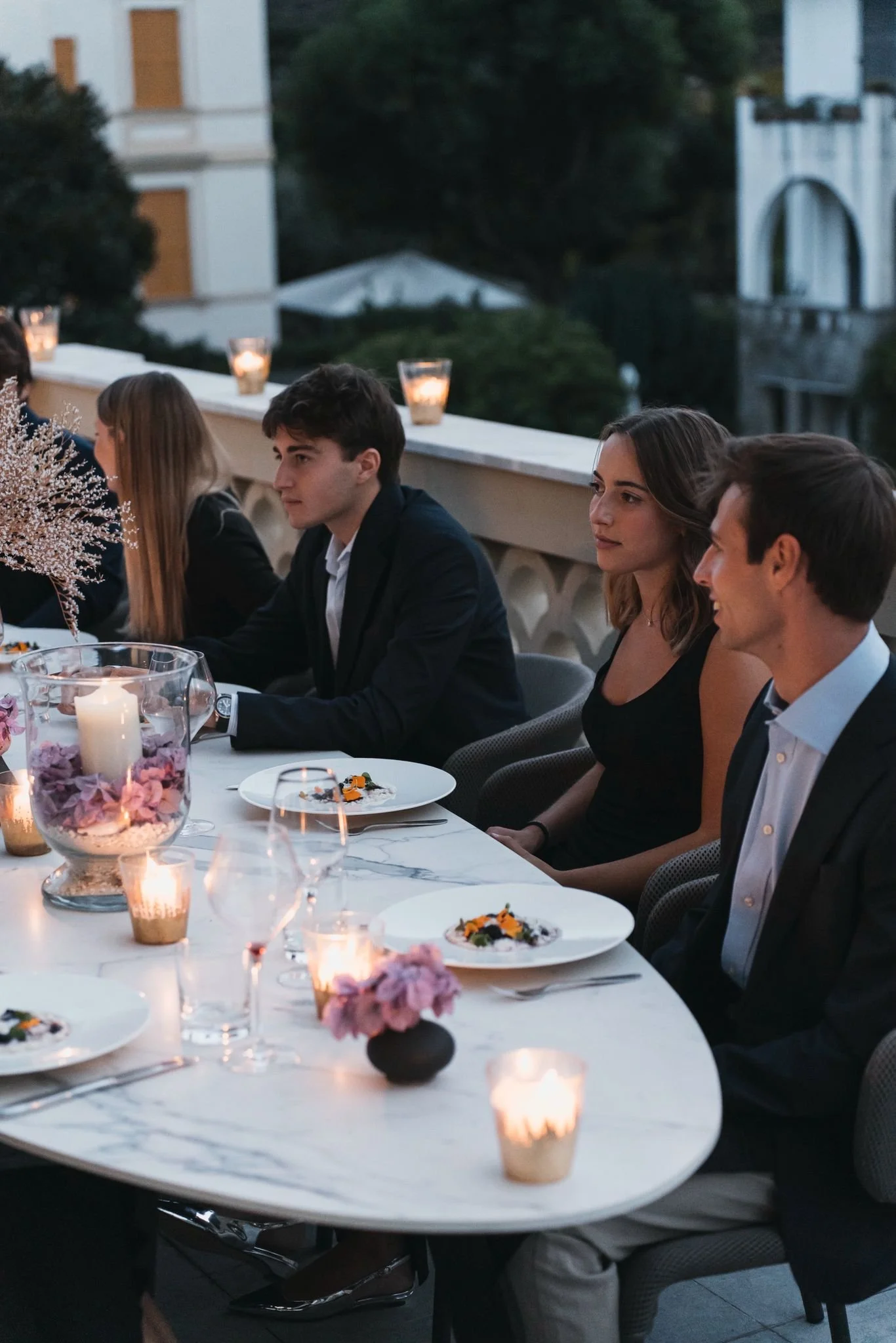 People sitting at a formal outdoor dinner table with lit candles, floral arrangements, and plated food, during evening or night.