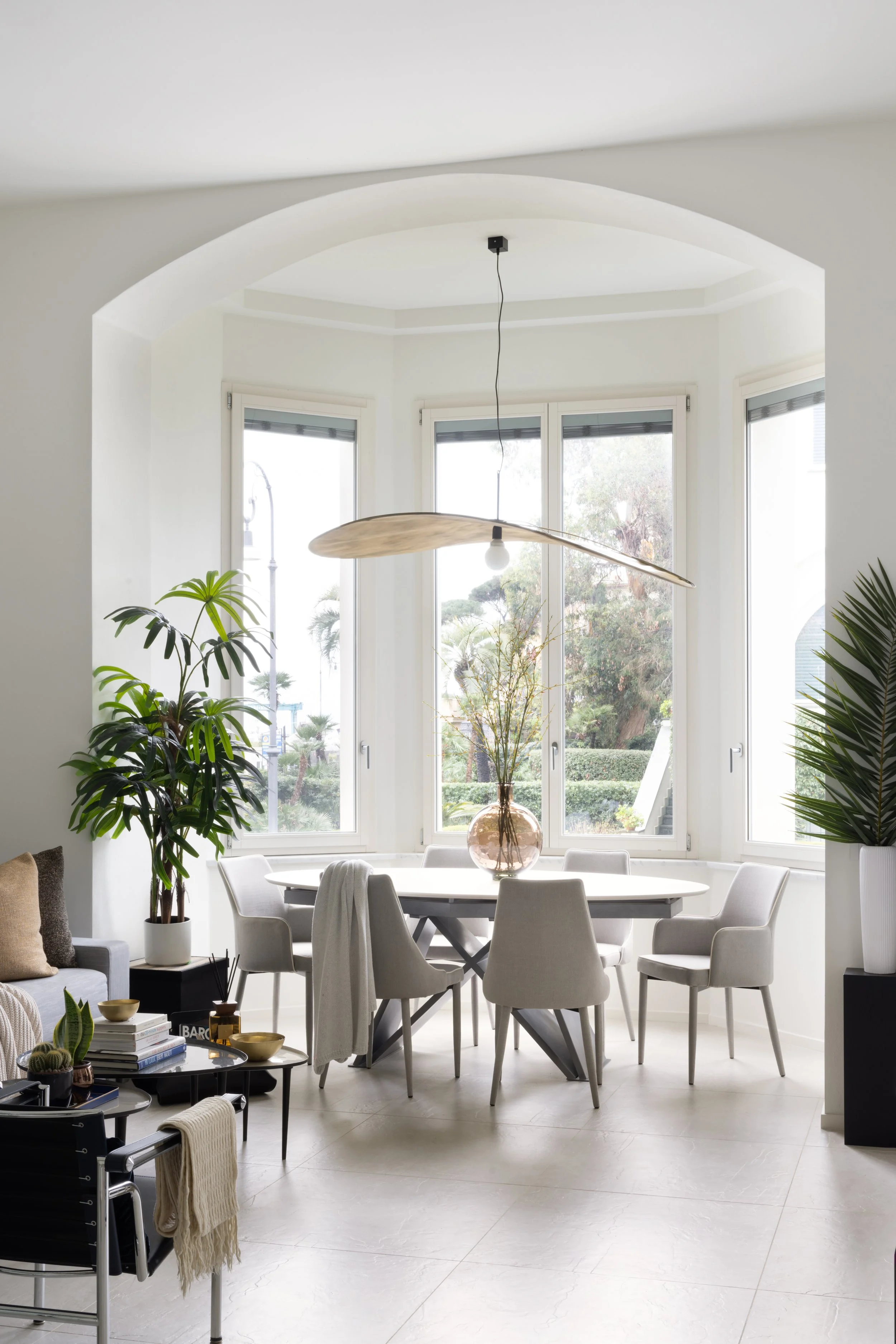 A modern dining room with a white oval table surrounded by eight white chairs. A large glass vase with tall plants is on the table. Large windows with black frames let in natural light, overlooking a garden with trees. There are plants in pots around