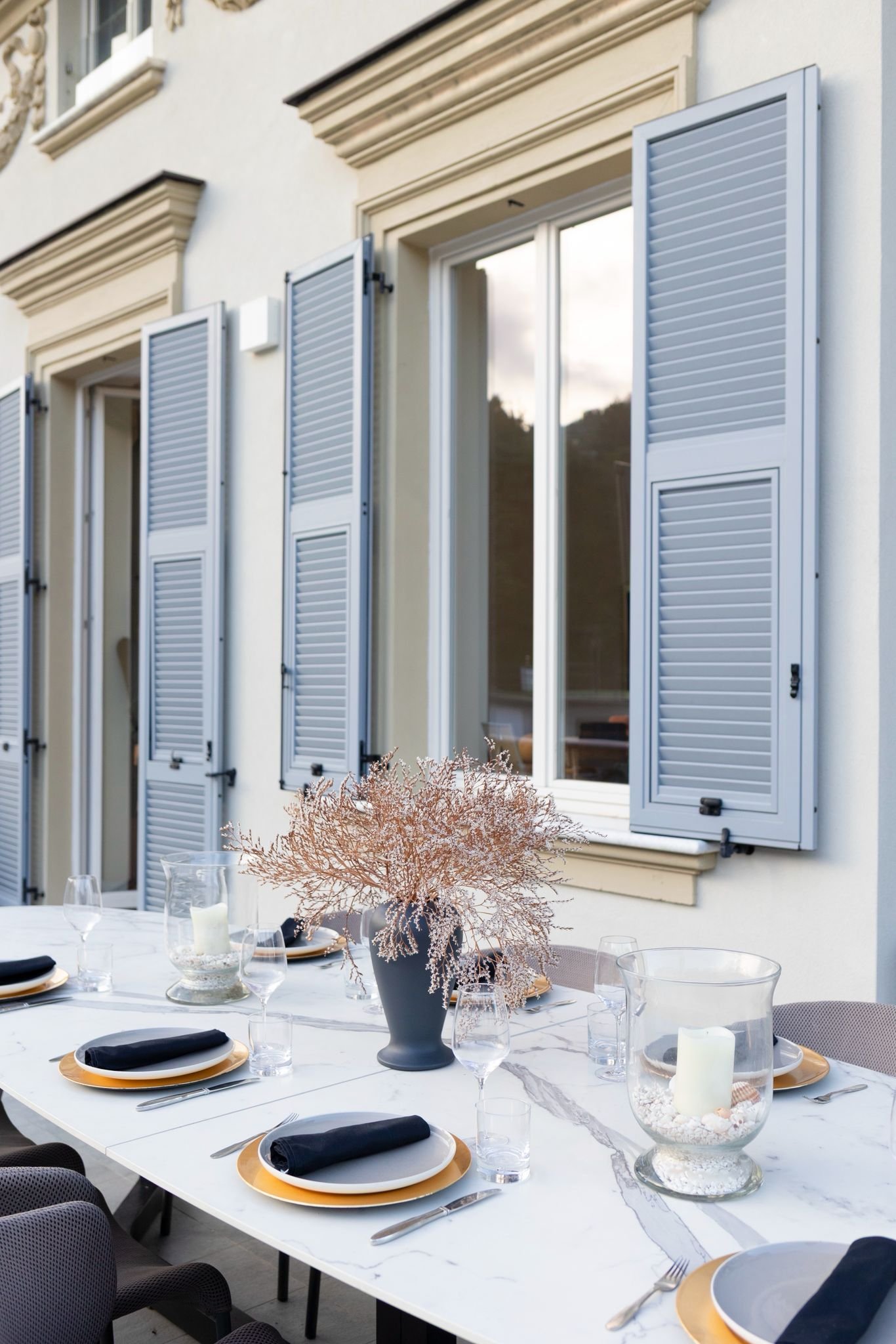 Outdoor dining table set with white and gold plates, black napkins, clear glasses, decorative centerpiece with pinkish branches in a dark vase, and candles in glass holders, in front of a building with large windows and light gray shutters.
