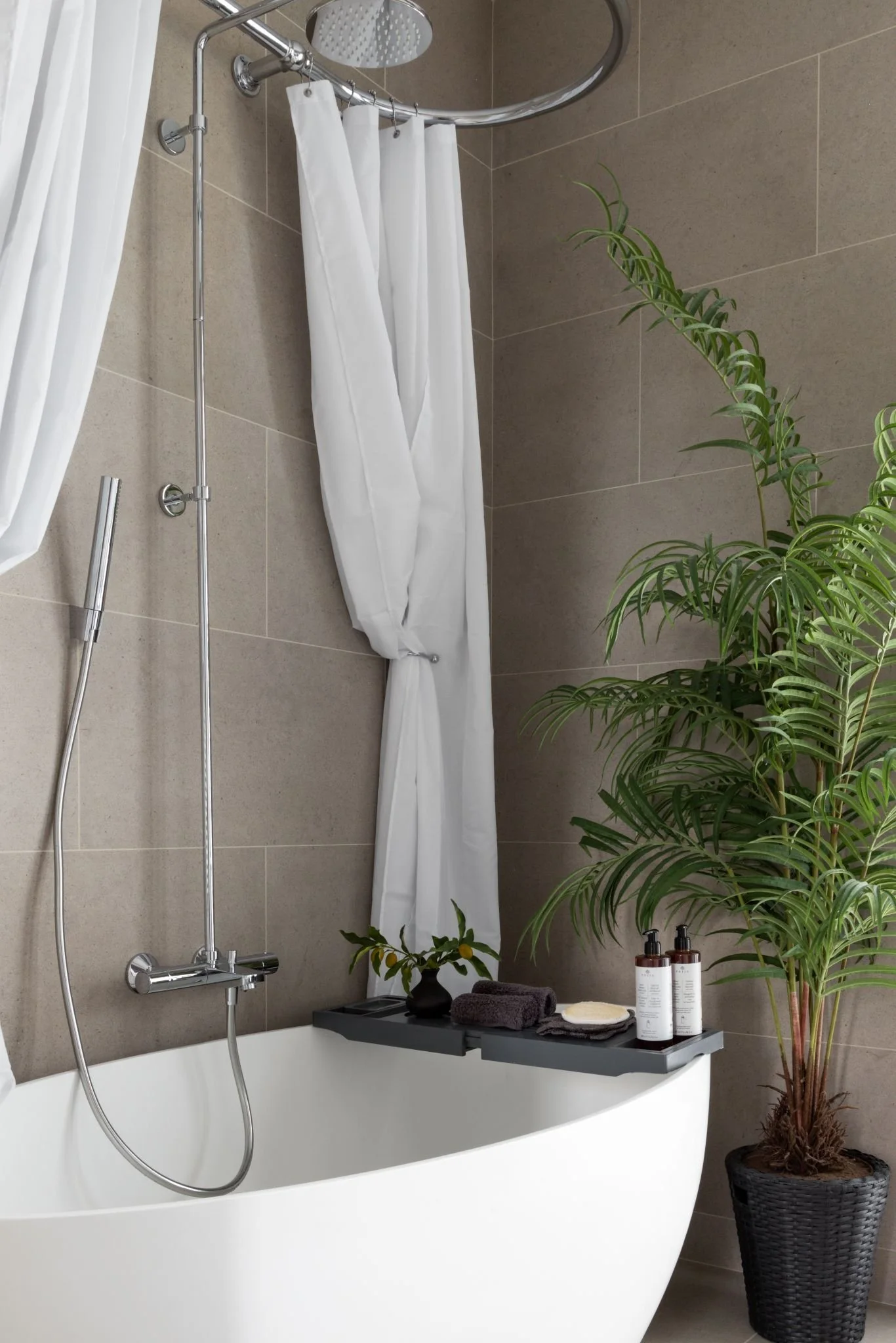 Modern bathroom with a white bathtub, shower area with curtain, a large potted plant, and a shelf with black towels and skincare products.
