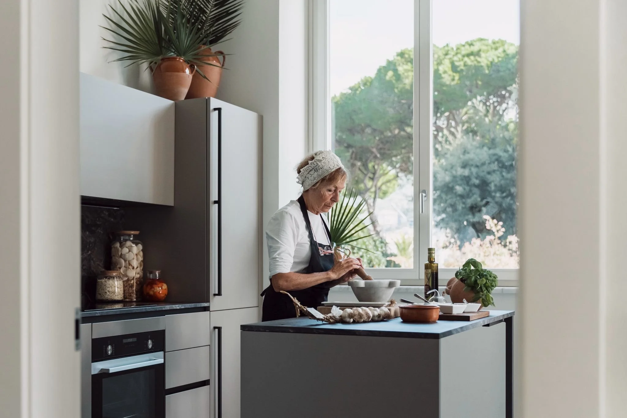 A woman in a kitchen chopping vegetables near a window with a view of trees outside.