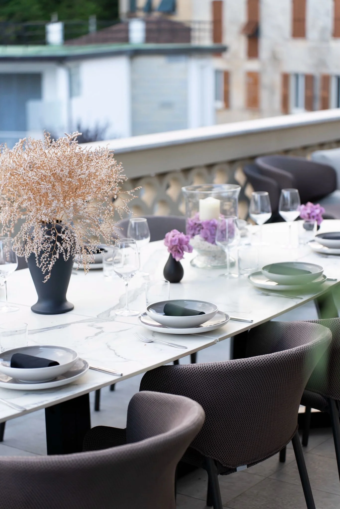 A dining table set for a meal on a balcony with floral centerpiece, candles, wine glasses, and plates, overlooking neighboring buildings.