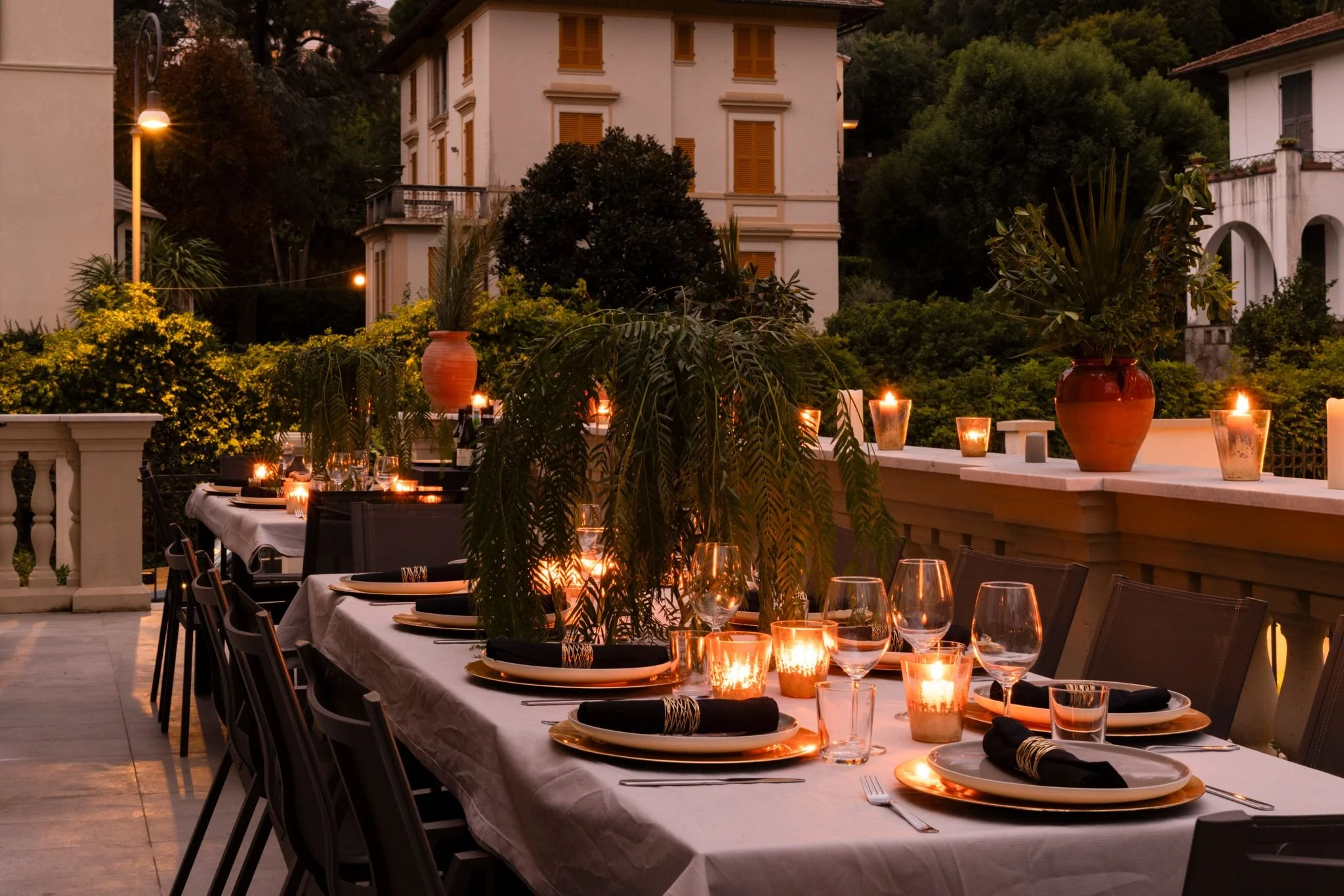 An outdoor dinner party at dusk with a long table set with black napkins, plates, wine glasses, and lit candles. There are large potted plants and a view of a house with brown shutters and lush greenery in the background.