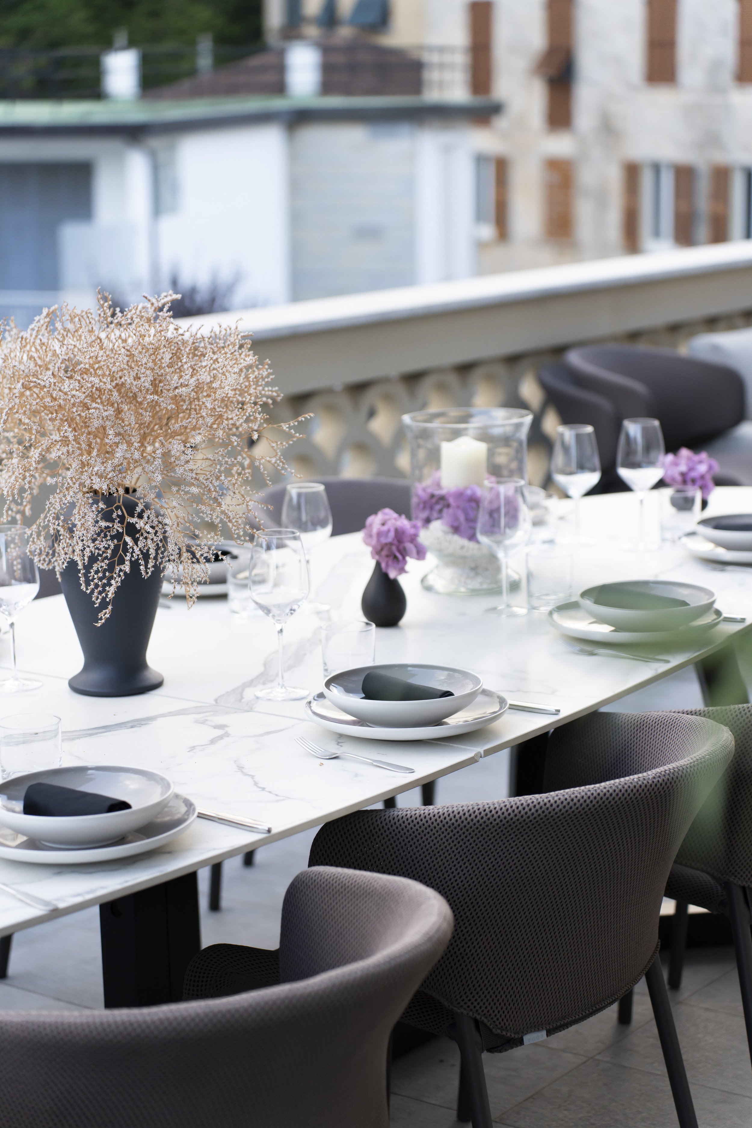 Dining table set for a meal with a large pink flower arrangement in a black vase, candles, wine glasses, and tableware, on a balcony with a cityscape background.