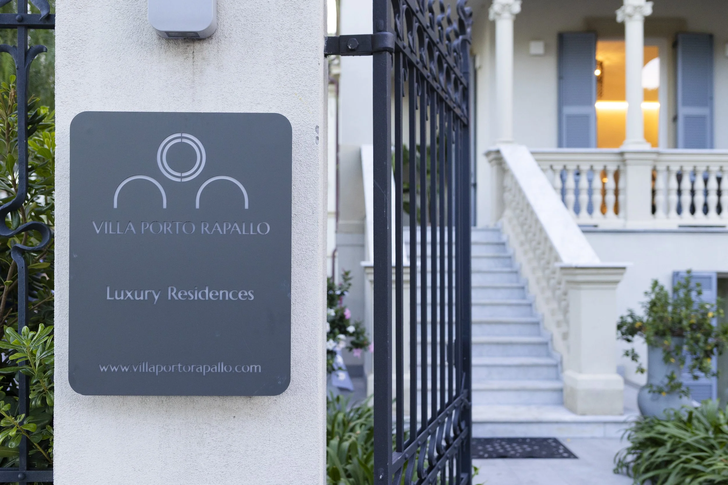 Entrance gate to Villa Porto Rapallo luxury residences, featuring a sign on a white wall and stairs leading up to the house with columns and potted plants.