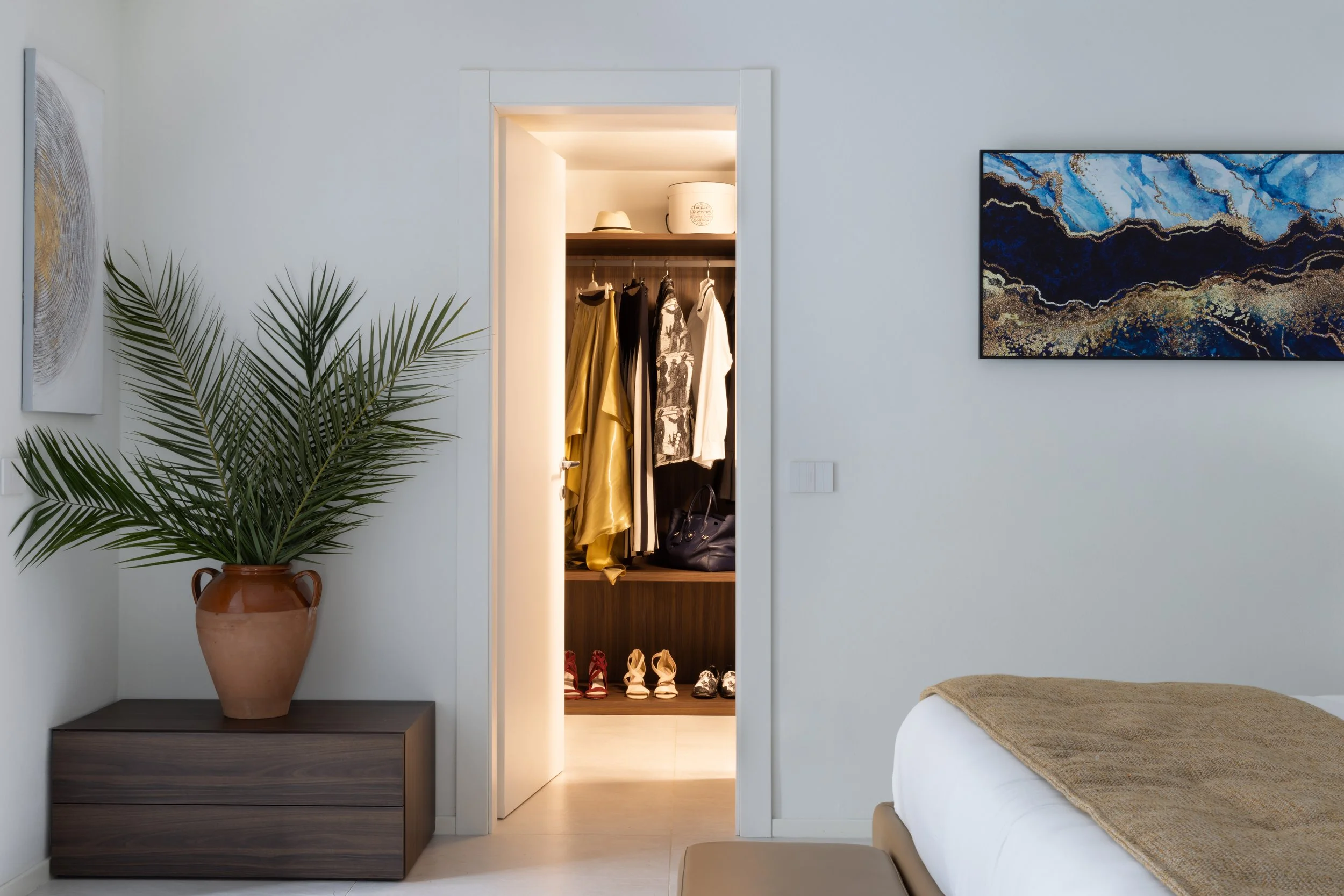 A bedroom with a large potted plant on a dark wooden nightstand, a built-in closet with hanging clothes and shoes inside, a large abstract blue and gold painting on the wall, and part of a bed with a beige blanket.