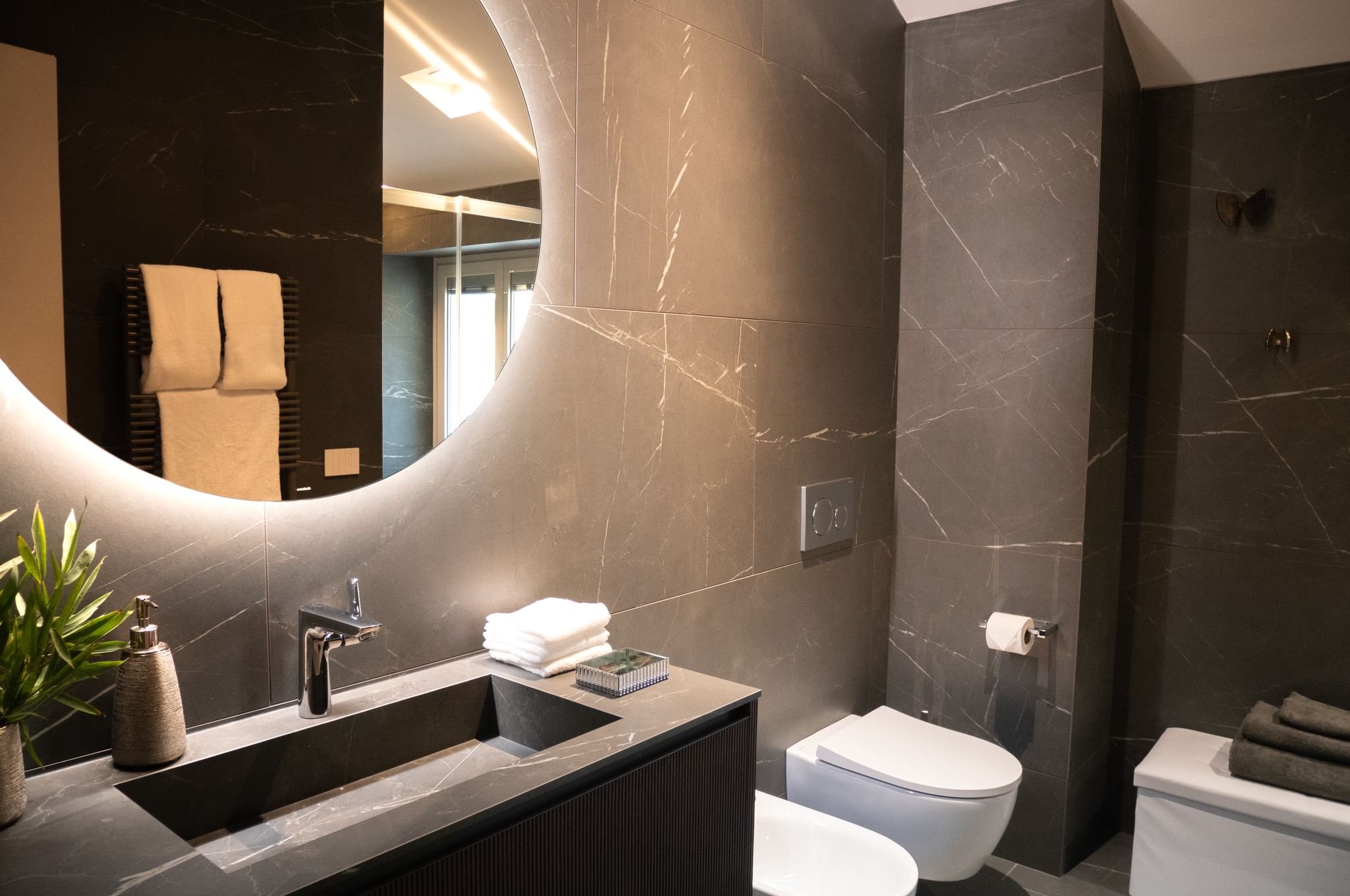 Modern bathroom with dark gray marble walls and countertop, large round mirror, white towels on a black rack, potted plant, and modern fixtures including a toilet, bidet, and sink.