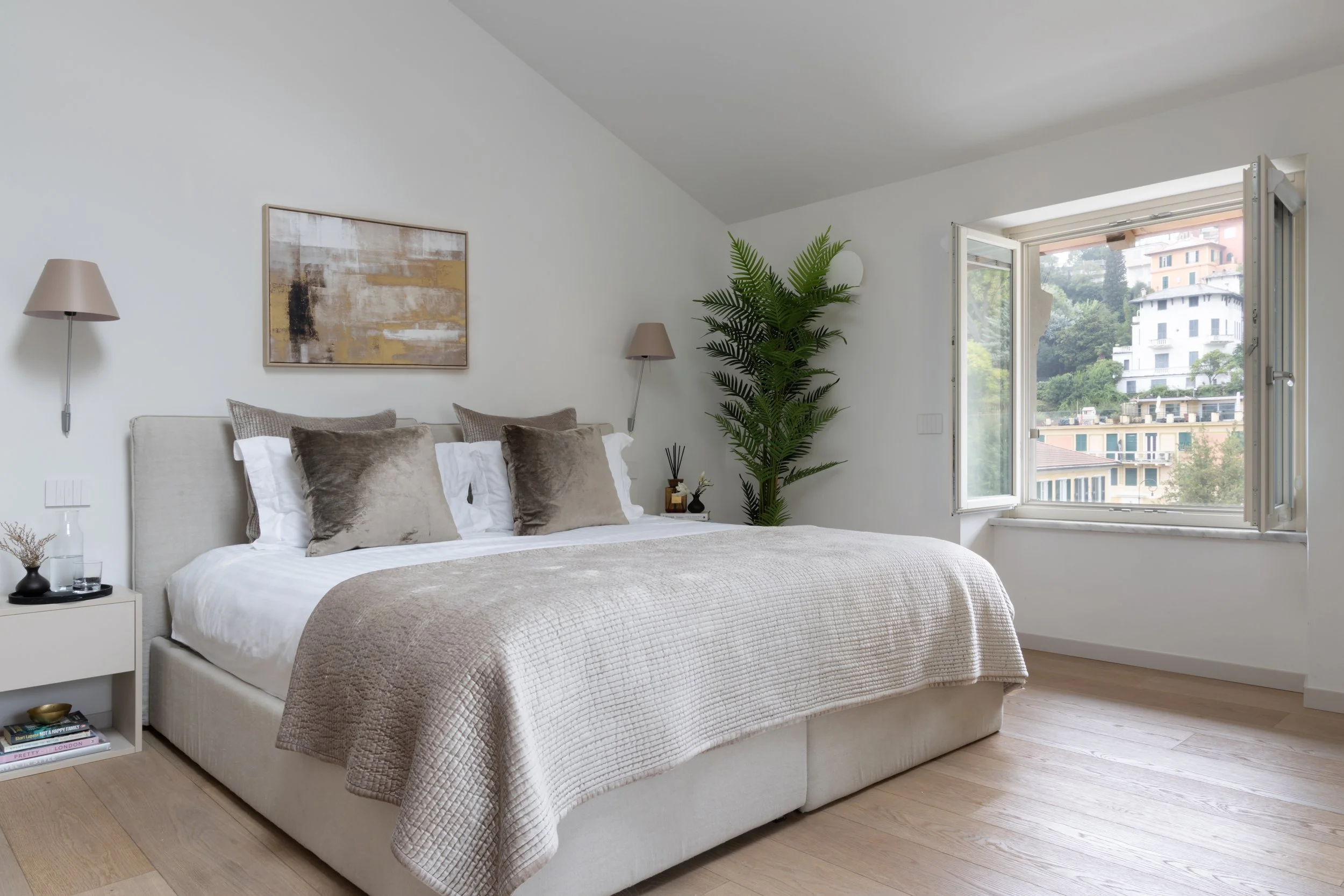 A cozy bedroom with a large bed featuring a beige quilt and multiple pillows, a white nightstand with books and a small decorative item, two wall-mounted lamps, a large abstract painting above the bed, a tall green plant near a window, and a view of 