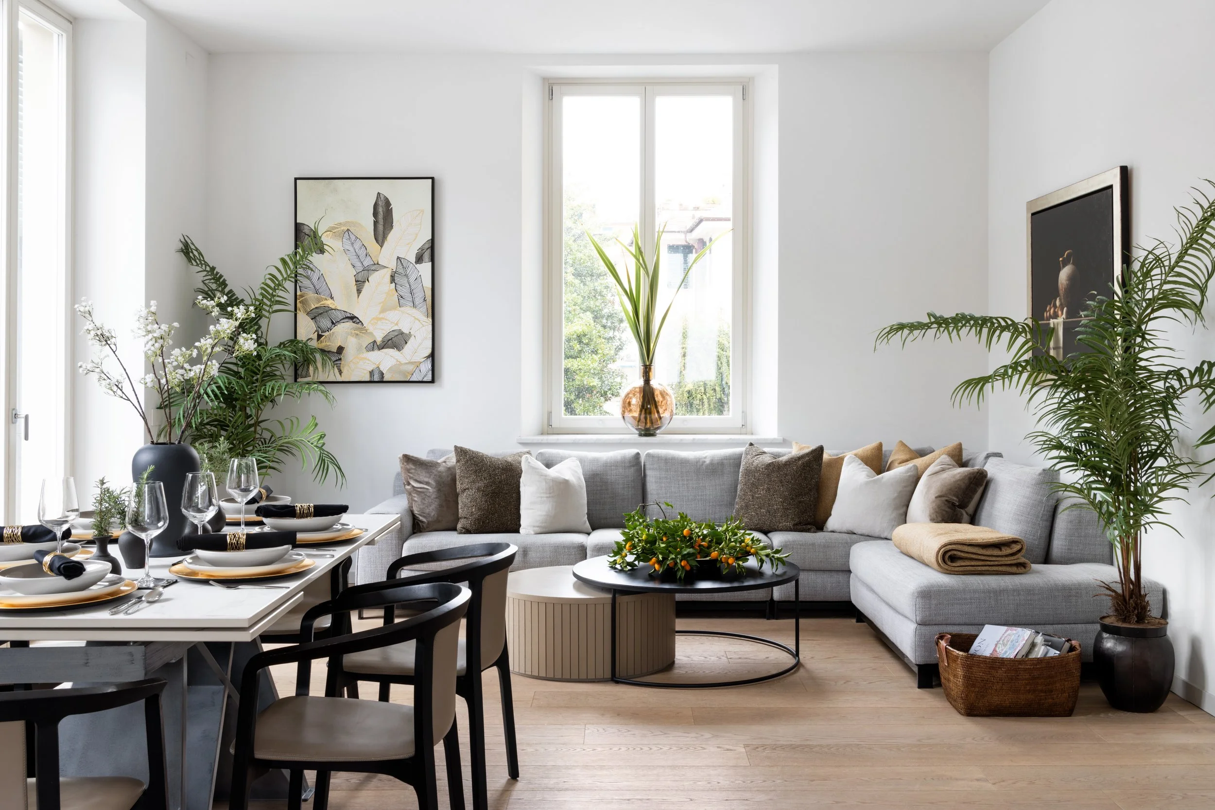 A bright dining and living room area with white walls, large windows, a sectional sofa, various plants, framed artwork, a dining table set for a meal, and cheerful modern decor.
