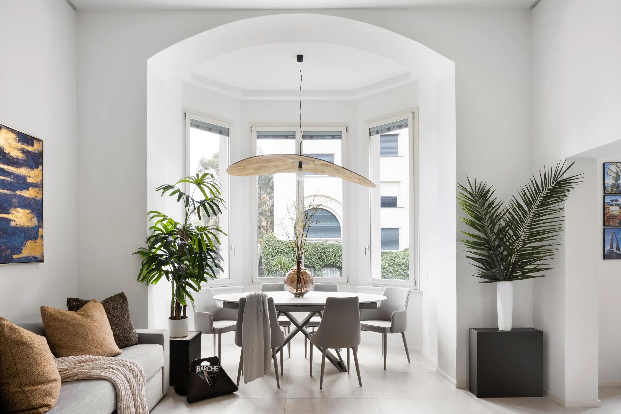 A modern dining room with large bay windows, a round dining table with chairs, potted plants, and wall art, with minimalist decor and natural light.