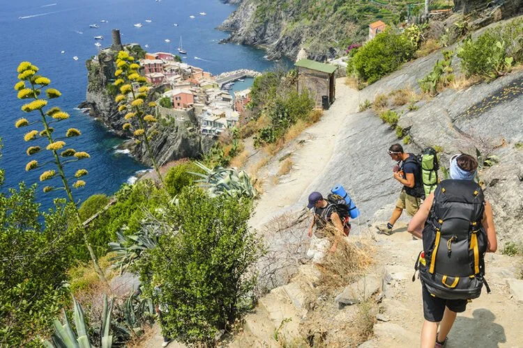 Three hikers with backpacks walking on a rocky trail along a hillside overlooking a coastal town with colorful buildings and a harbor, with the ocean in the background.