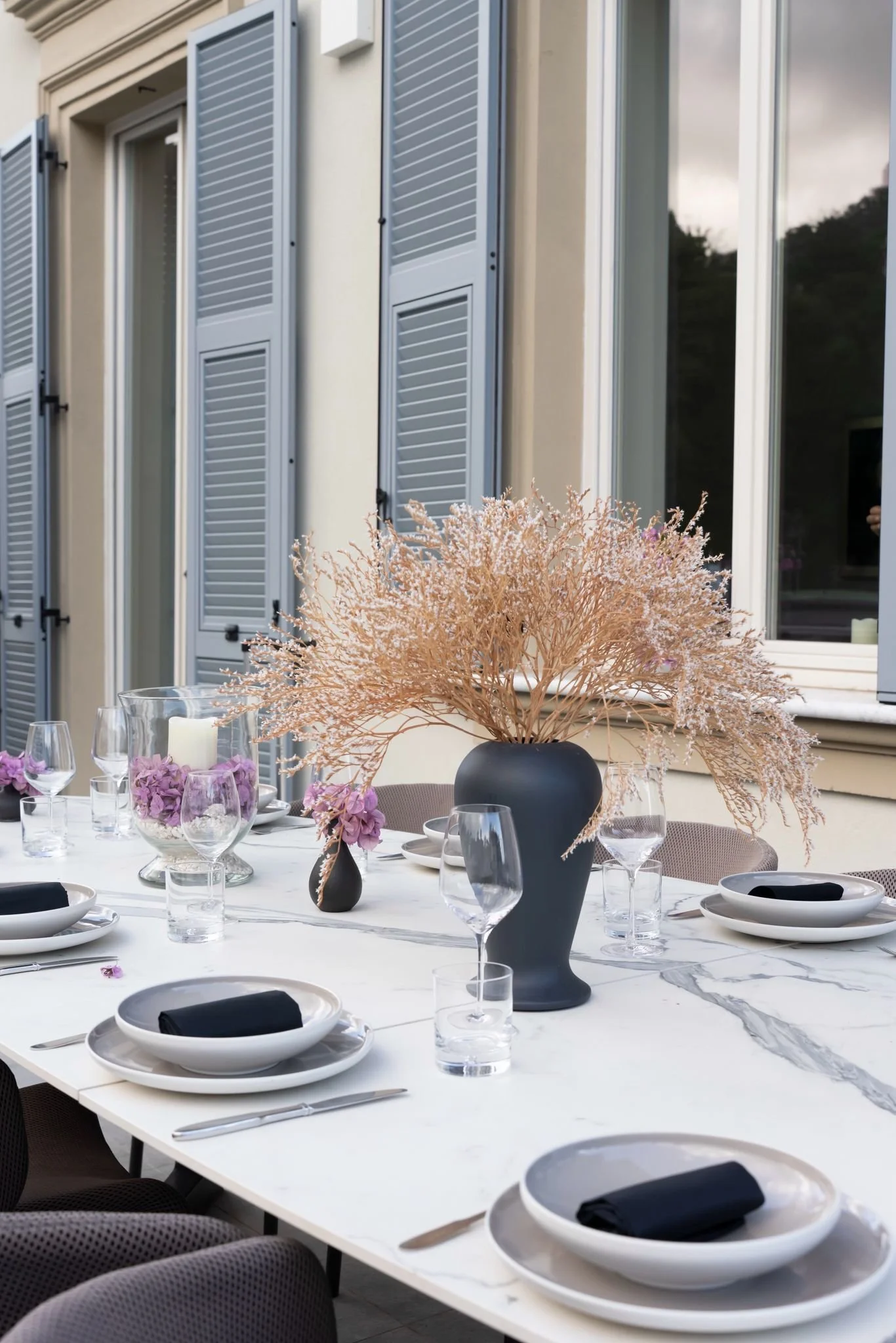A dining table set outdoors with black and white tableware, black napkins, and clear glasses, decorated with tall pinkish-beige dried flowers in black vases, in front of a house with white walls and blue shutters.
