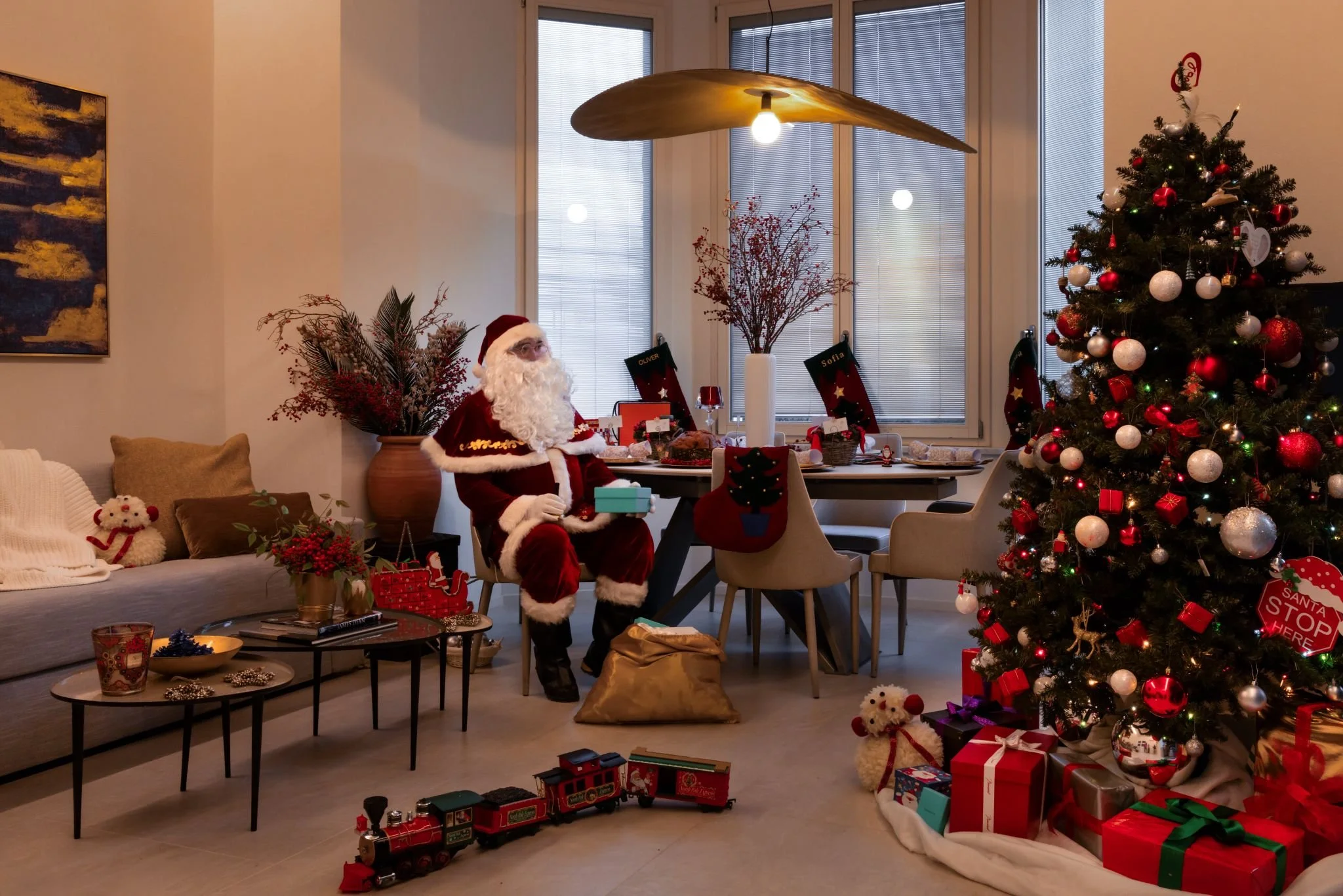 Living room decorated for Christmas with a large decorated Christmas tree, a Santa Claus figure sitting near a table, wrapped presents, and holiday decorations, including stockings hanging on the window.