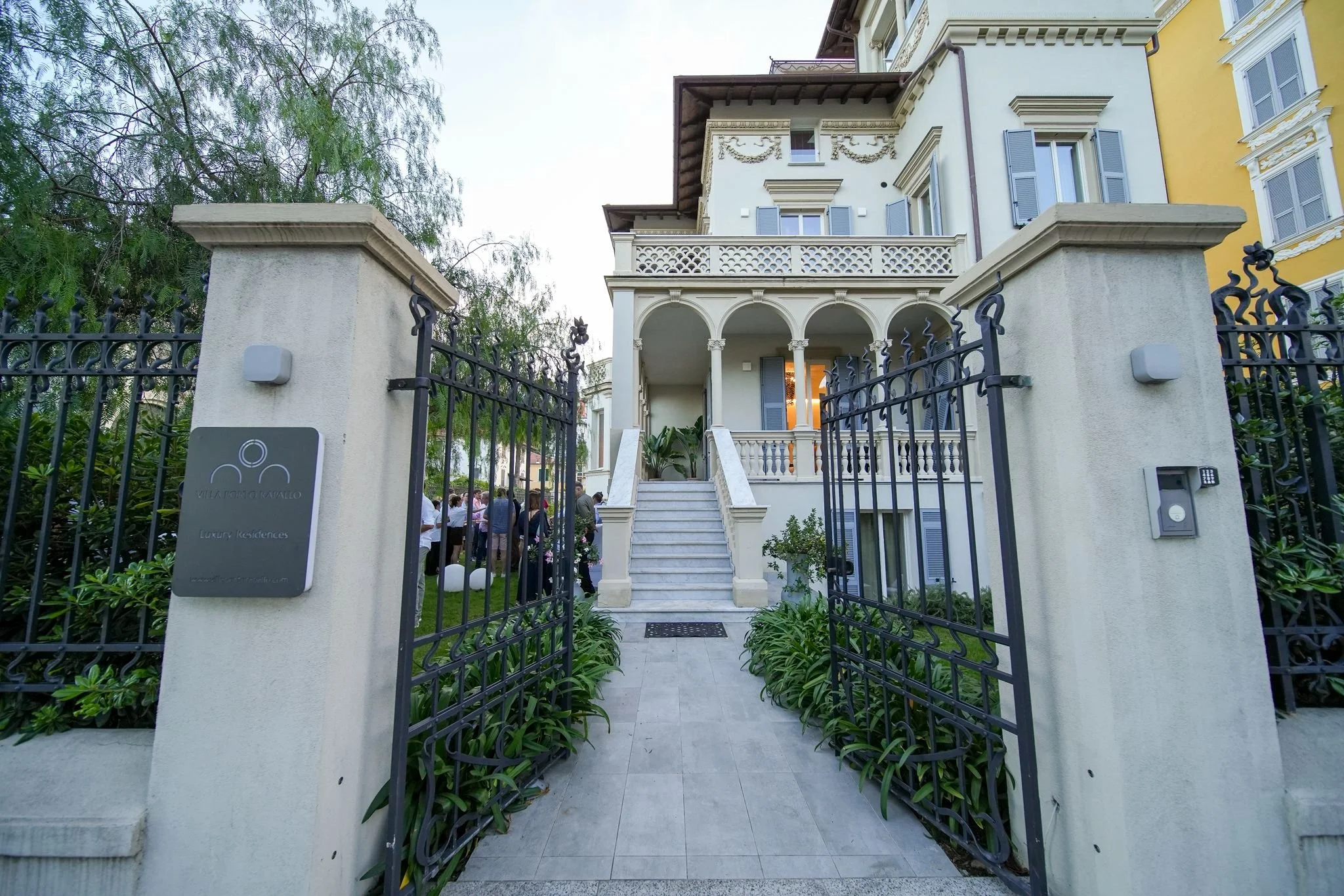 A gated entrance leading to a white multi-story building with classic architectural details, stairs, and a small garden with plants, with a group of people standing in front of the building.