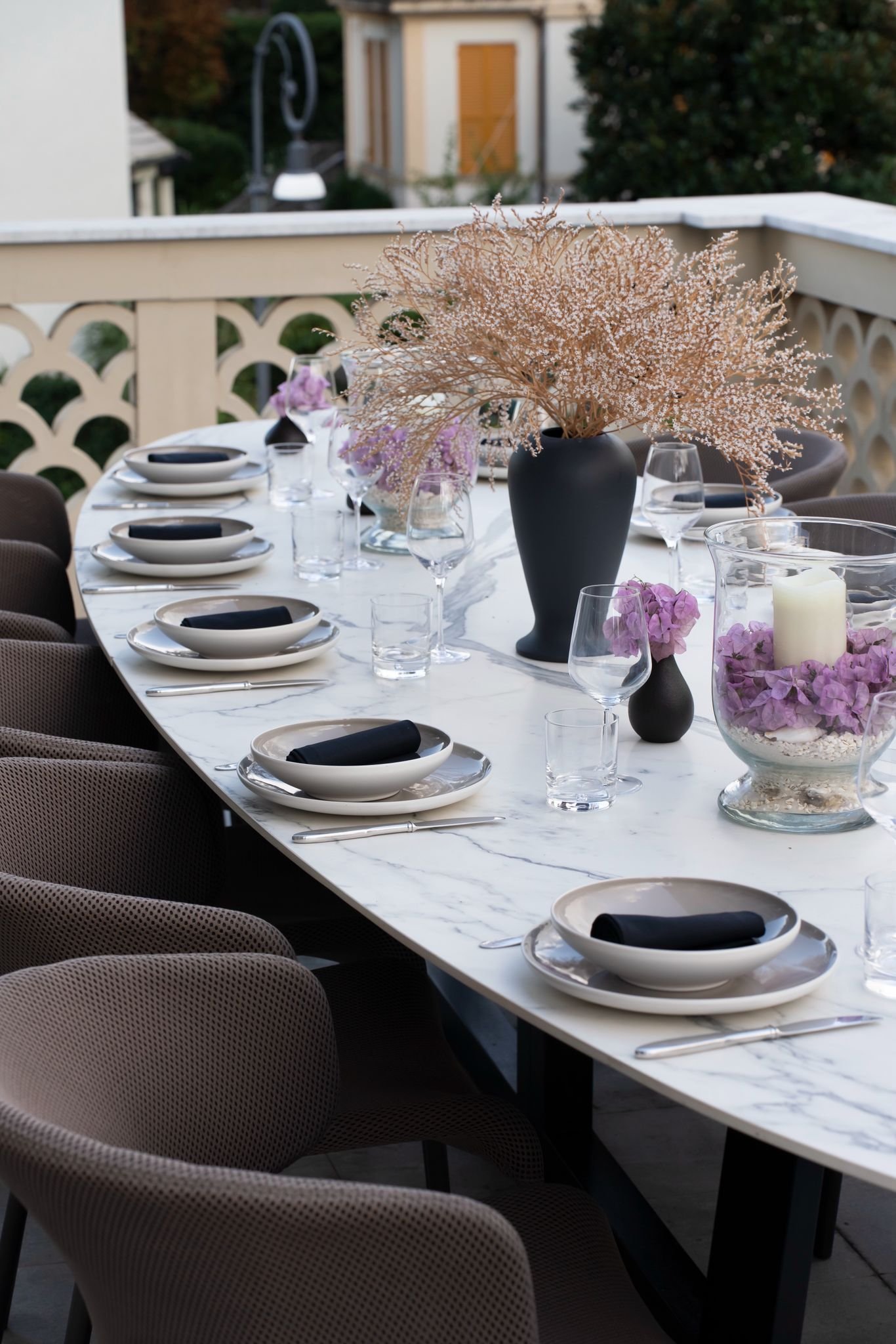 A decorated outdoor dining table with plates, glasses, and black napkins, featuring a large black vase with pink and white flowers, set on a marble surface on a balcony with a decorative railing, surrounded by plants and trees.