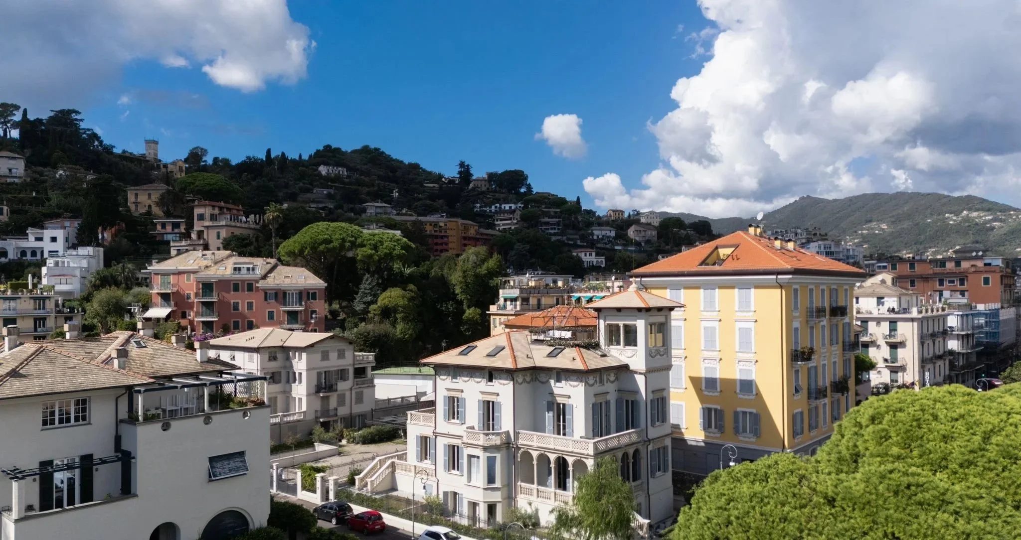 A scenic view of a hillside city with multicolored buildings, lush green trees, and a partly cloudy sky.