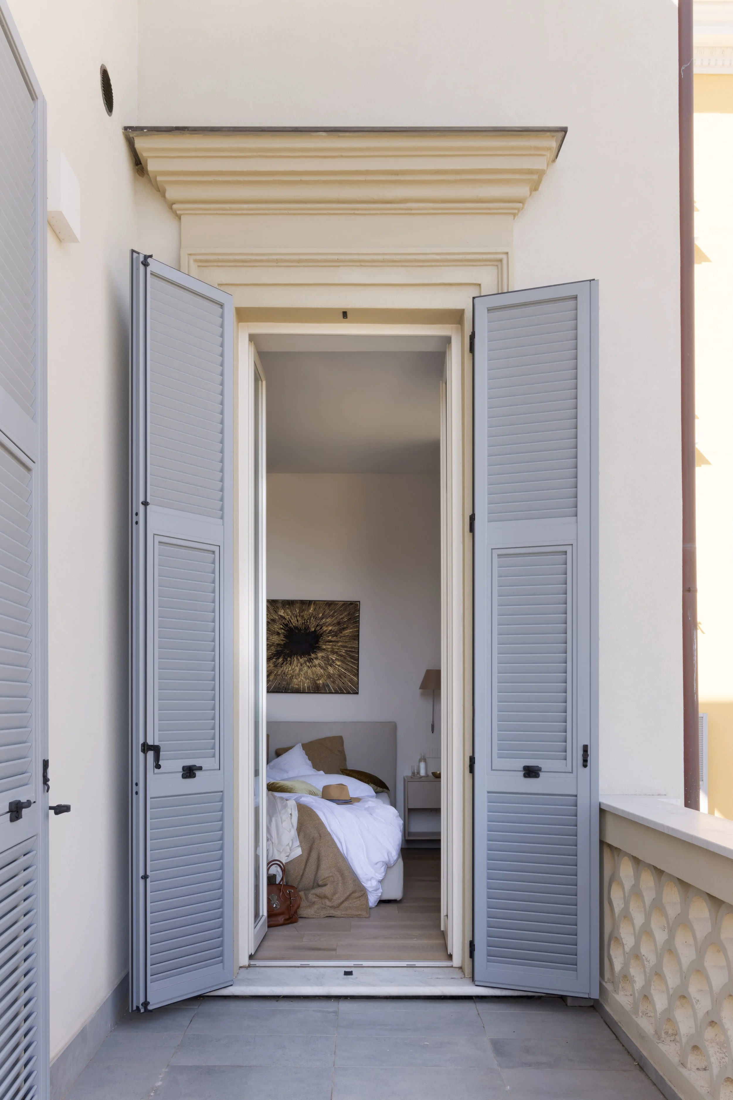 Bedroom visible through open French doors with gray shutters, a bed with white linens, tan blanket, pillows, bedside table, and artwork on the wall.