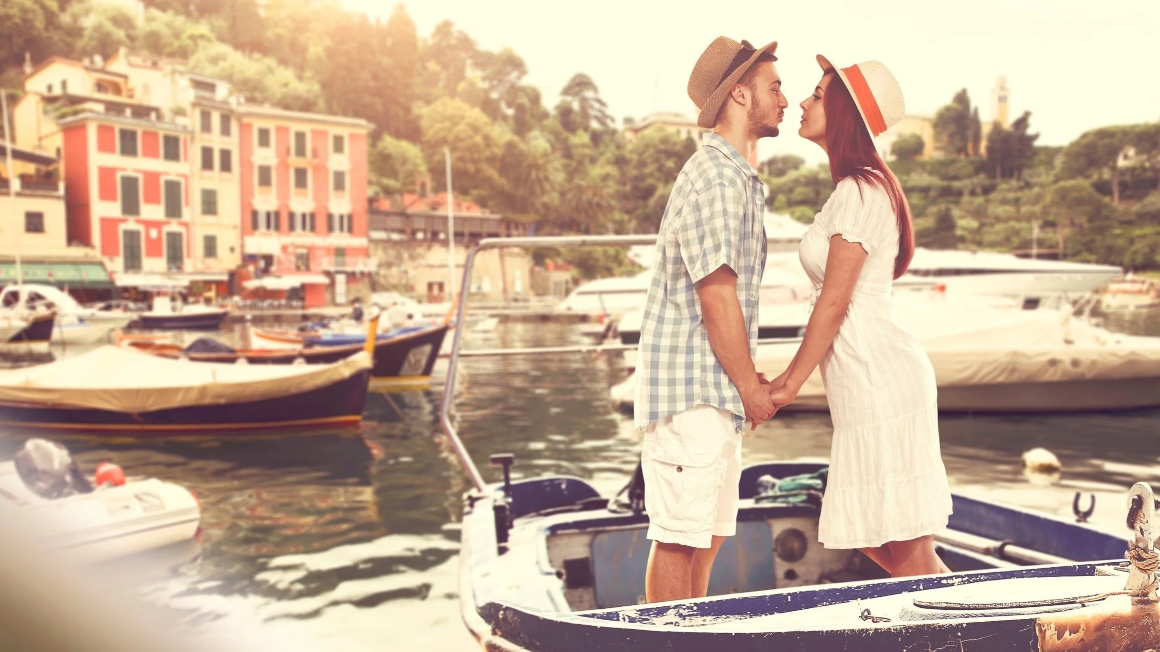 A couple holding hands and about to kiss on a boat in a harbor with colorful buildings and trees in the background.
