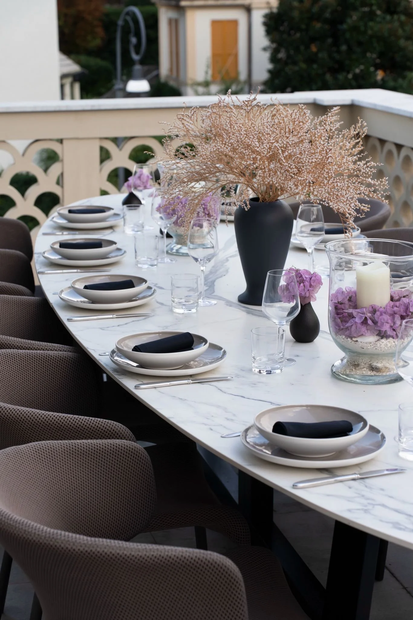 A decorated outdoor dining table with a large black vase holding pink and white flowers as a centerpiece, set with black napkins, white plates, wine glasses, and candles, surrounded by beige chairs, overlooking a white railing and greenery in the bac