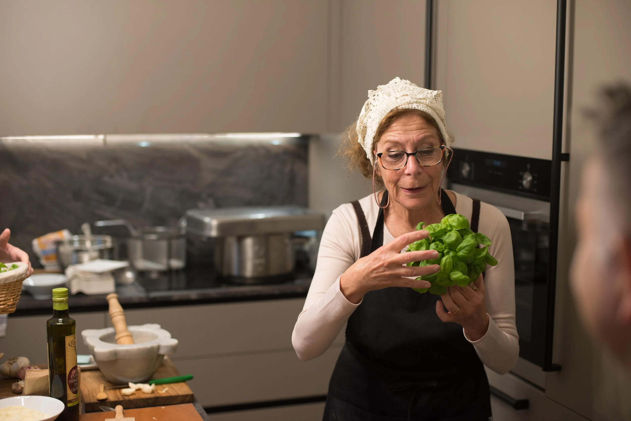 An older woman with glasses and a beige headscarf holding a bunch of fresh basil in a kitchen with stove and cooking ingredients on the counter.