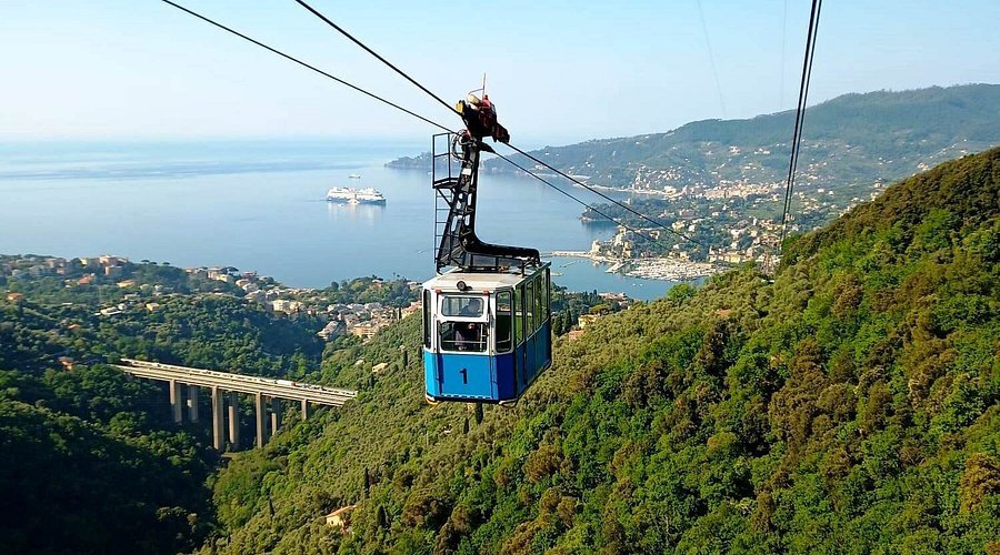 A blue and white cable car traveling over green hills with a lake and a small city in the background.