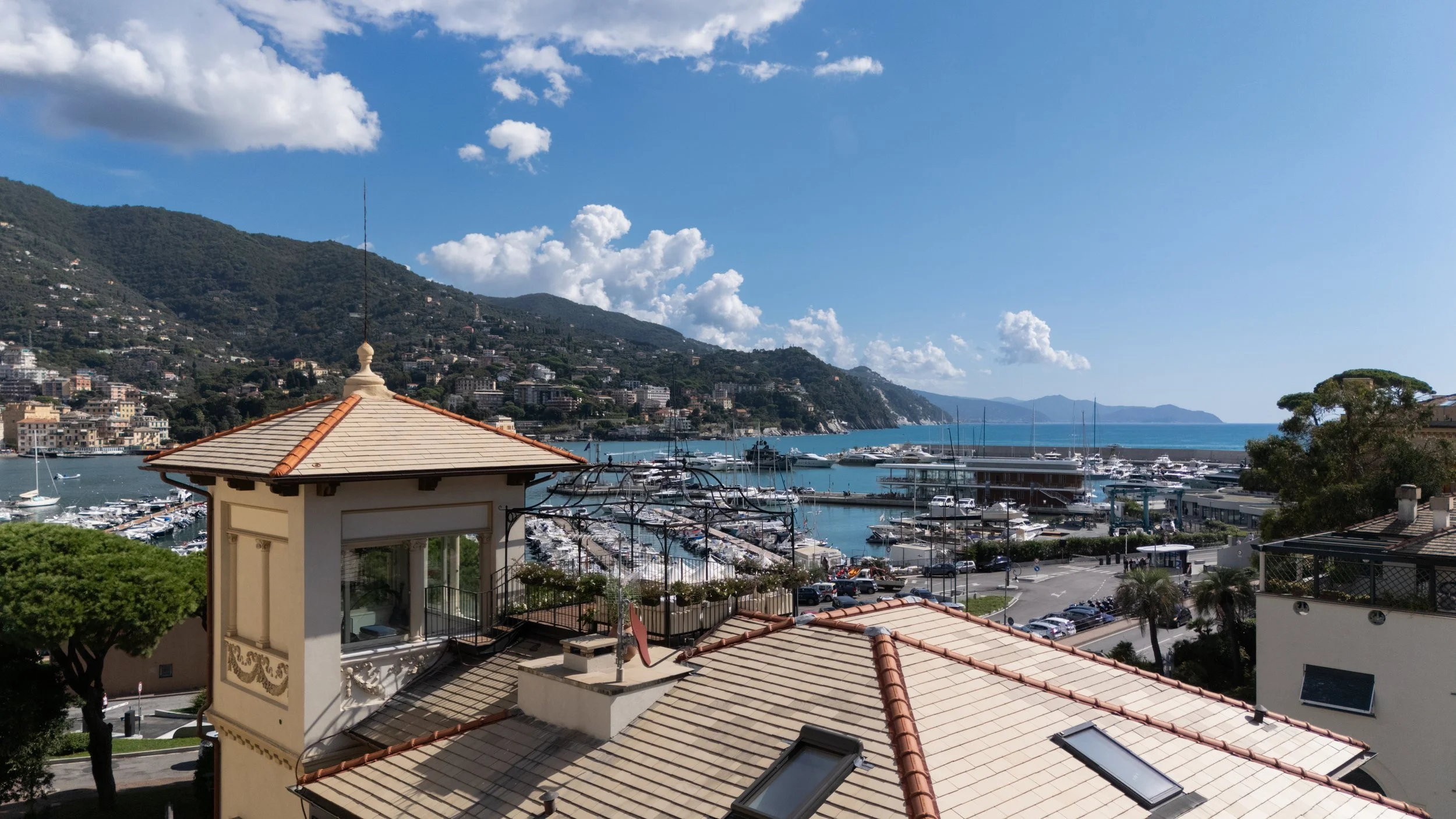 View of a marina with boats and yachts, surrounded by hills with buildings, under a partly cloudy sky.