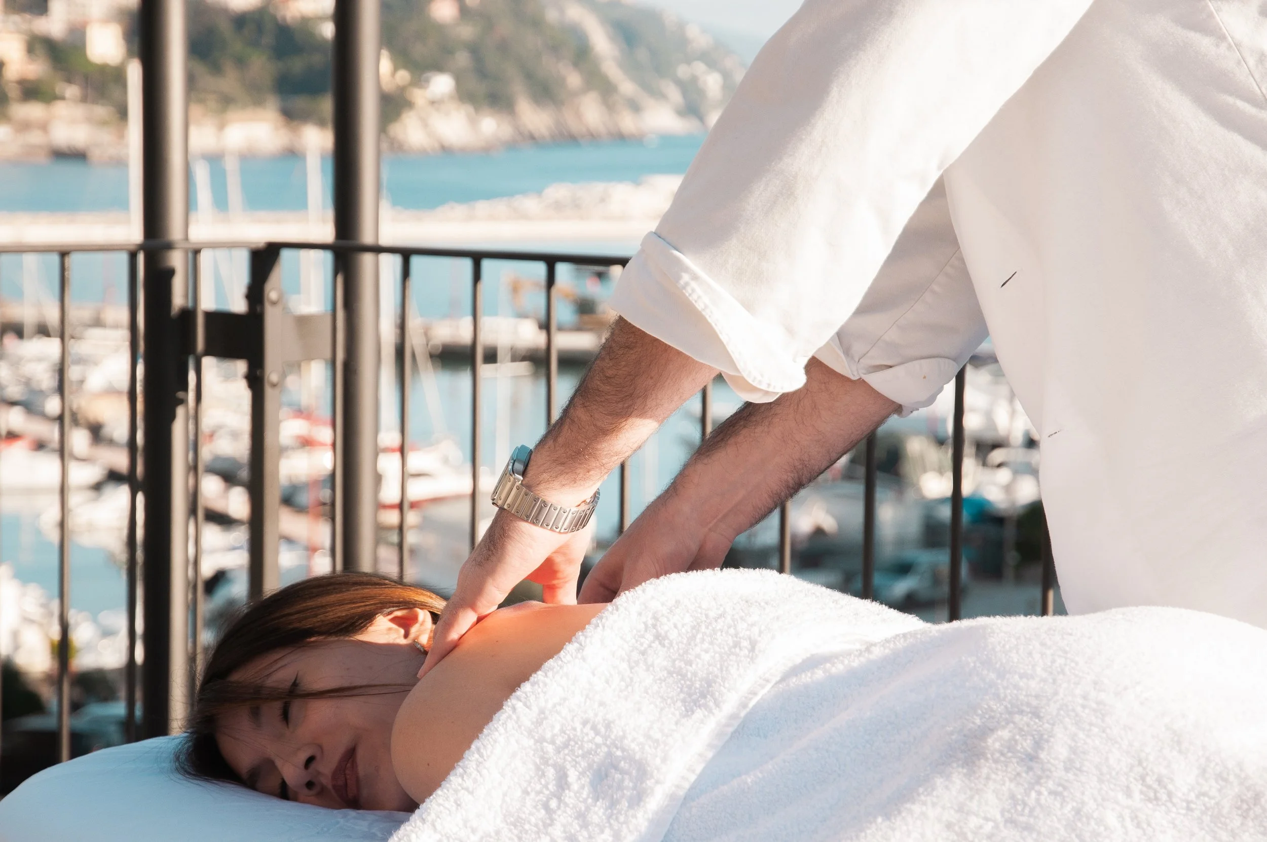 A woman receiving a massage outdoors near a marina, with a scenic view of water and boats in the background.