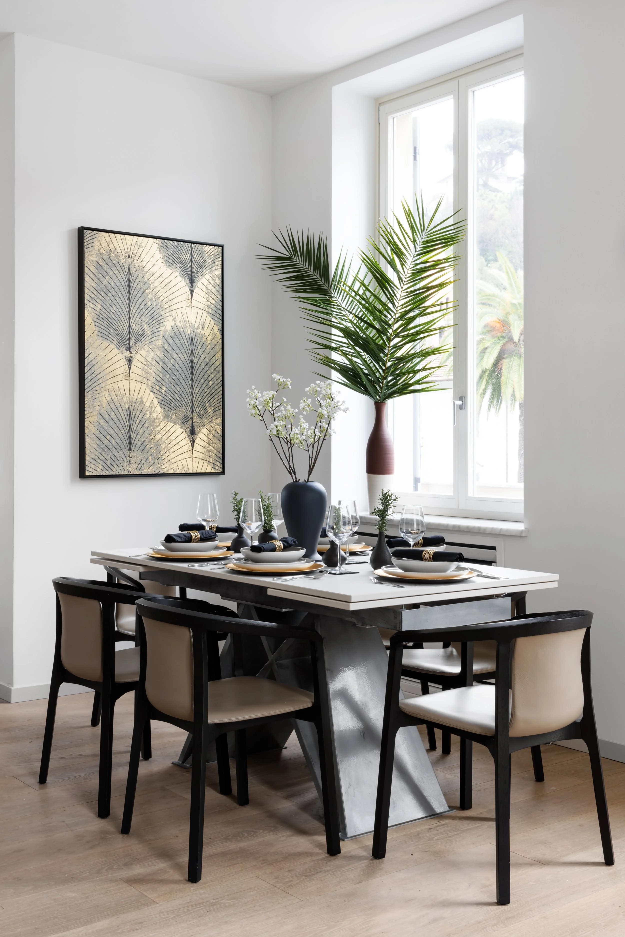 A modern dining room with a white table set for six, black chairs, and a black vase with white flowers. A large window shows outdoor palm trees. Artwork with a pattern of fan-shaped leaves hangs on the white wall.