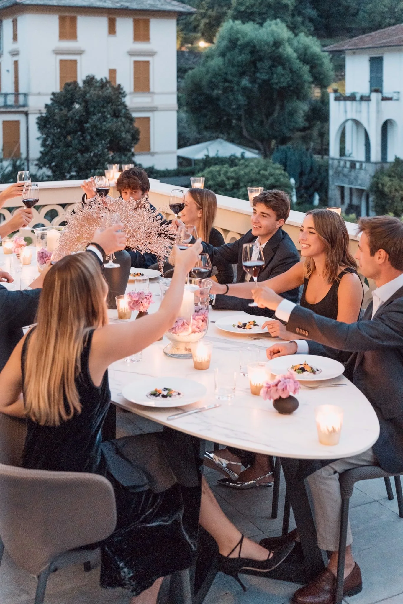 A group of well-dressed young adults celebrating at a dinner party on an outdoor terrace, raising glasses of red wine in toast, with a decorated table, candles, and some pink floral arrangements, overlooking a scenic residential area with trees and h