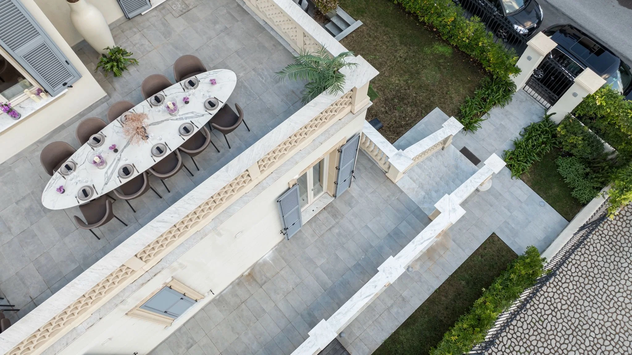 A rooftop outdoor dining area featuring an oval marble table with nine chairs, decorative candles, and flower arrangements, surrounded by railings and greenery.