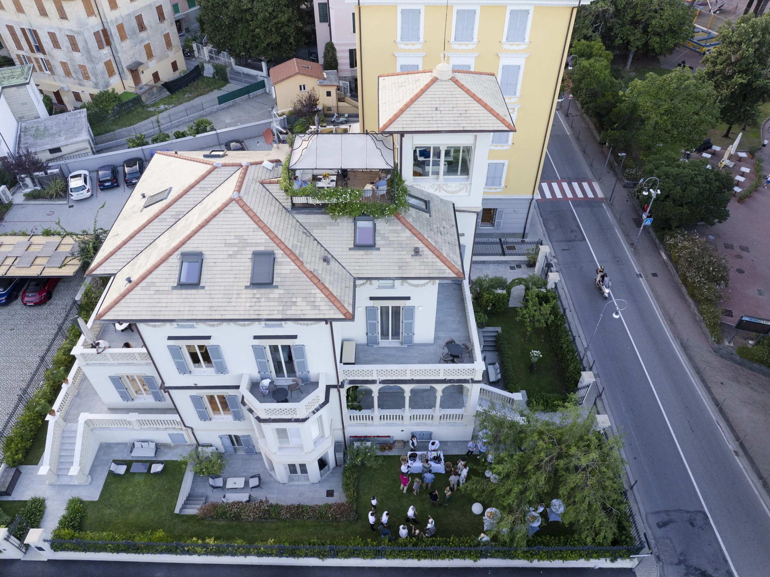 Aerial view of a white building with a rooftop terrace where several people are gathered. The surrounding area includes a small parking lot, greenery, trees, and a street with pedestrians and a cyclist.