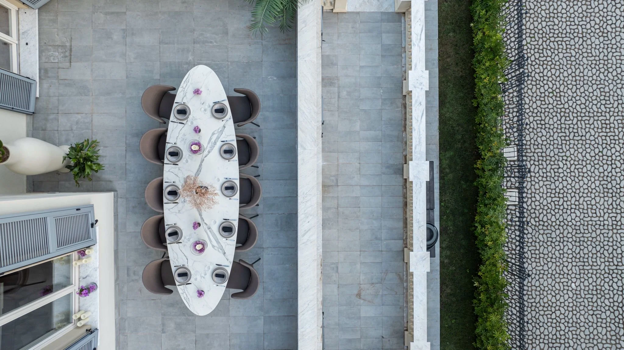 A top-down view of an outdoor patio area featuring a white marble dining table with six brown chairs around it. The table is decorated with purple flowers and candles. To the left, there is a large white vase with green plants, and the patio is surro