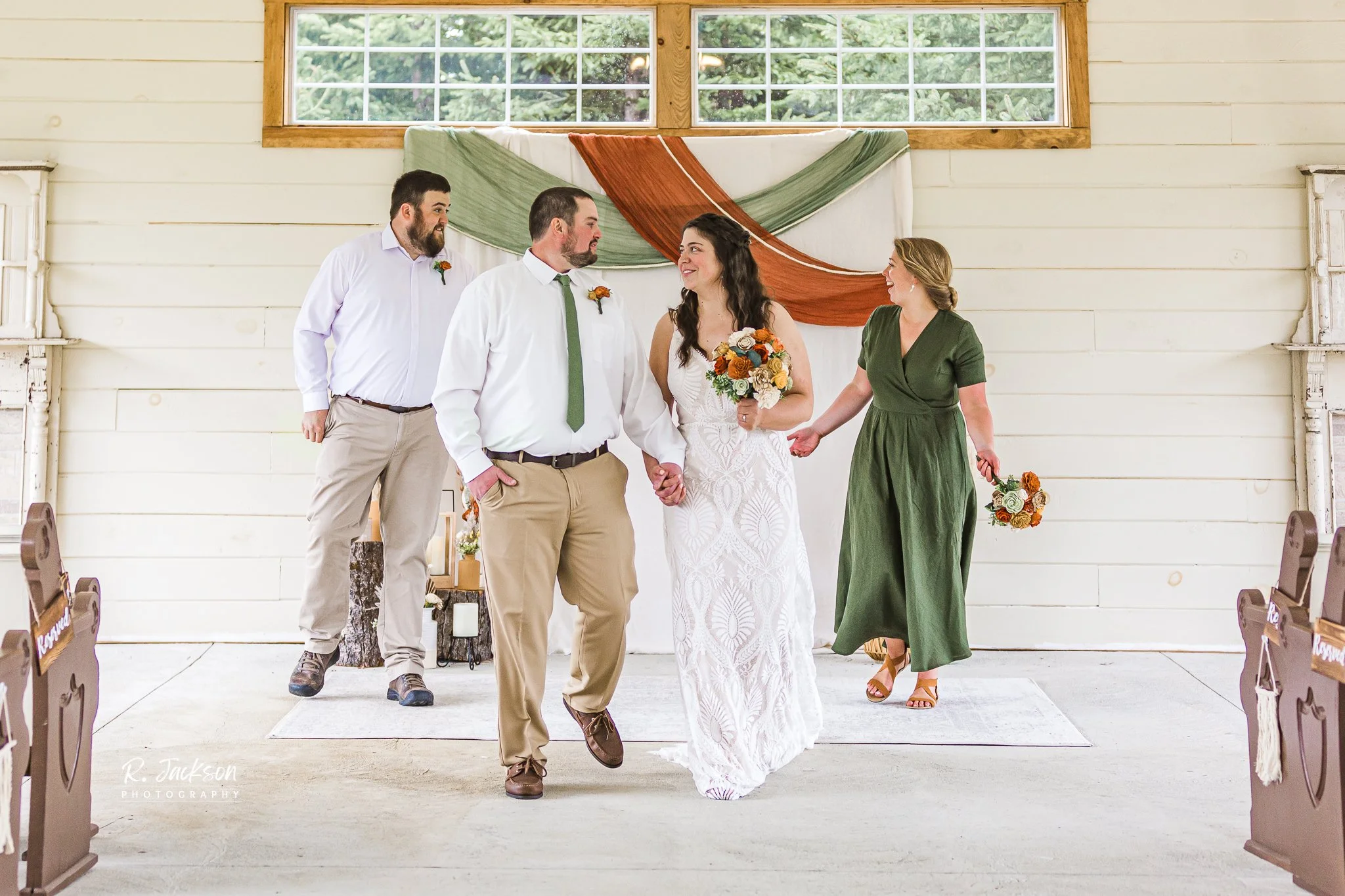 A couple gets married inside a rustic wedding venue with white wooden paneled walls and a large window. The bride holds a bouquet, and they are holding hands while walking together. The officiant is at the right, smiling and holding the bride's hand. Two men stand on the left, watching the ceremony. The decor features green and rust-colored fabric draped behind the couple and a small wooden table with candles.