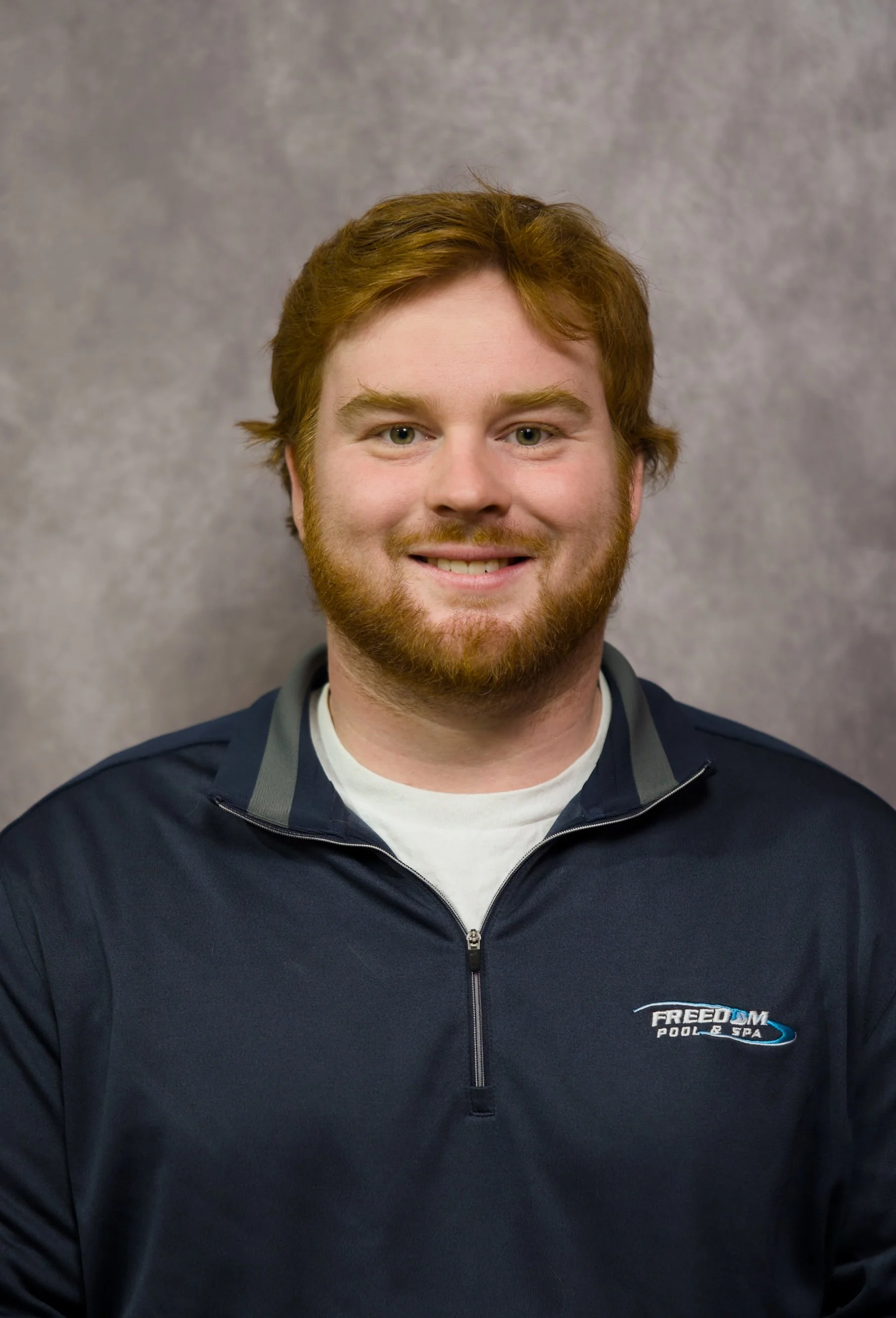 A young man with red hair and a beard smiling at the camera, wearing a navy blue quarter-zip jacket with 'FREEDOM Pool & Spa' embroidered on it, against a gray textured background.