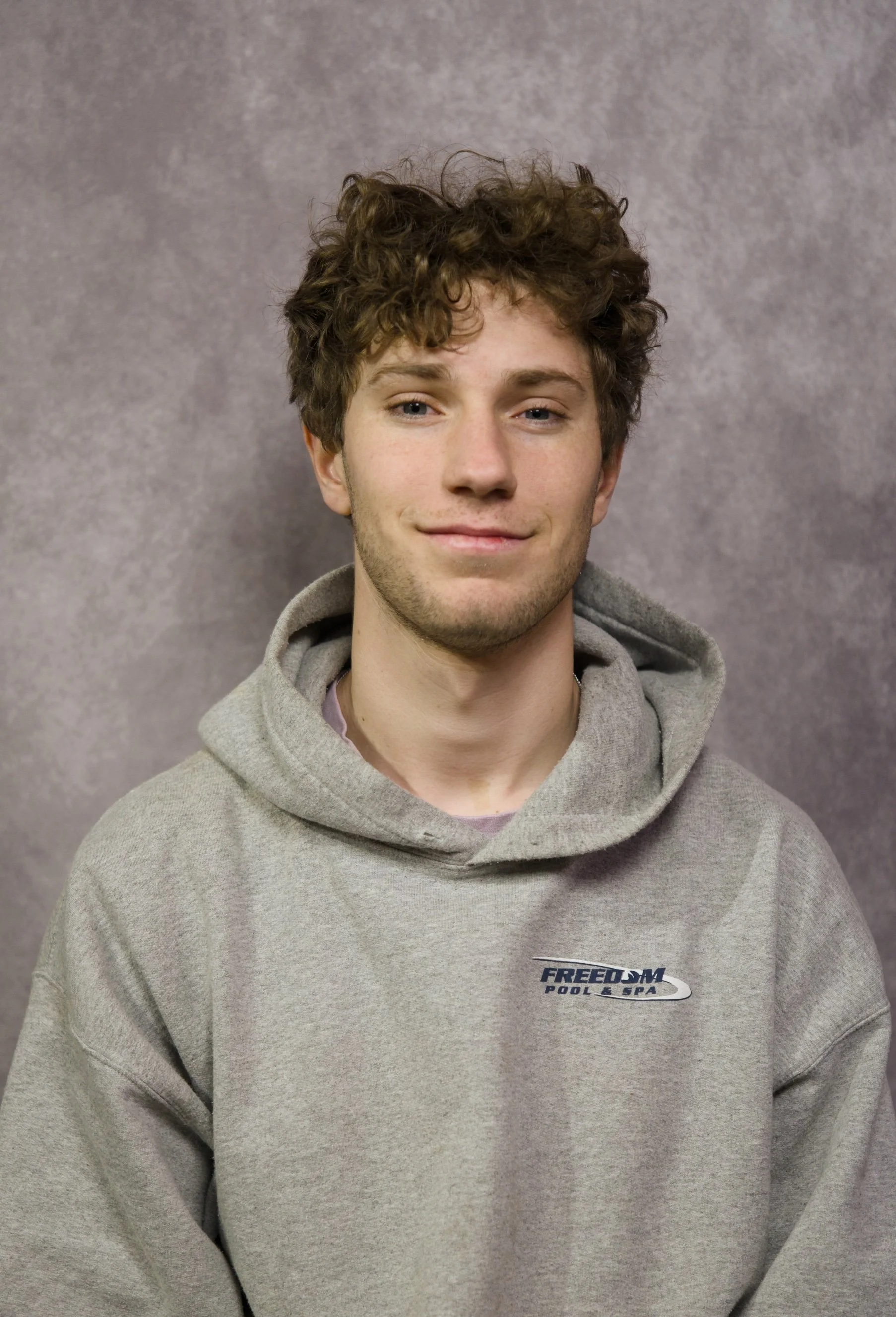 A young man with curly brown hair and light skin, wearing a gray hooded sweatshirt with a "FREEDOM Pool & Spa" logo, smiling slightly at the camera, standing against a mauve textured background.