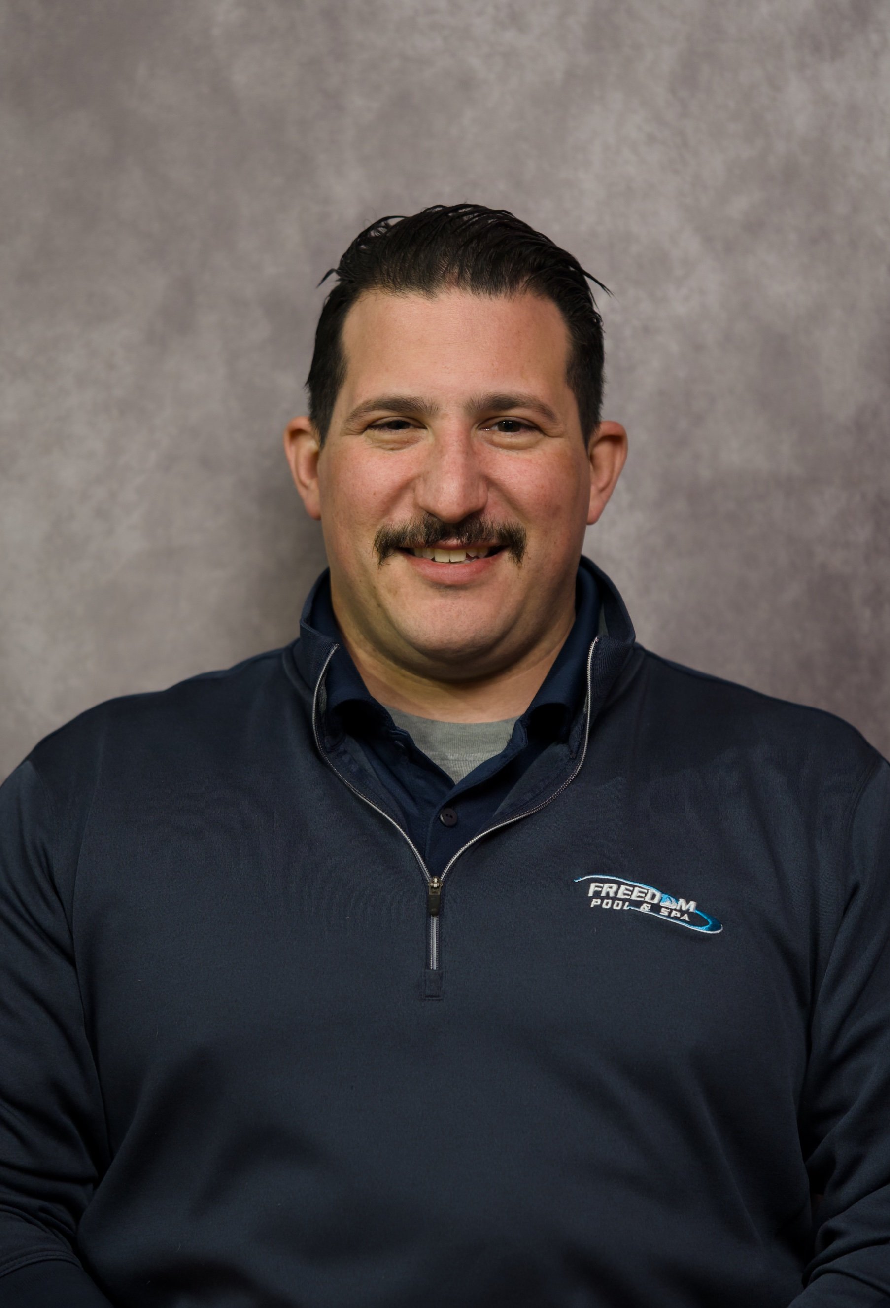 Portrait of a man with dark hair, mustache, and light skin, smiling, wearing a navy blue zip-up jacket with 'FREEDOM POOLS & SPA' logo, standing against a plain, gray background.