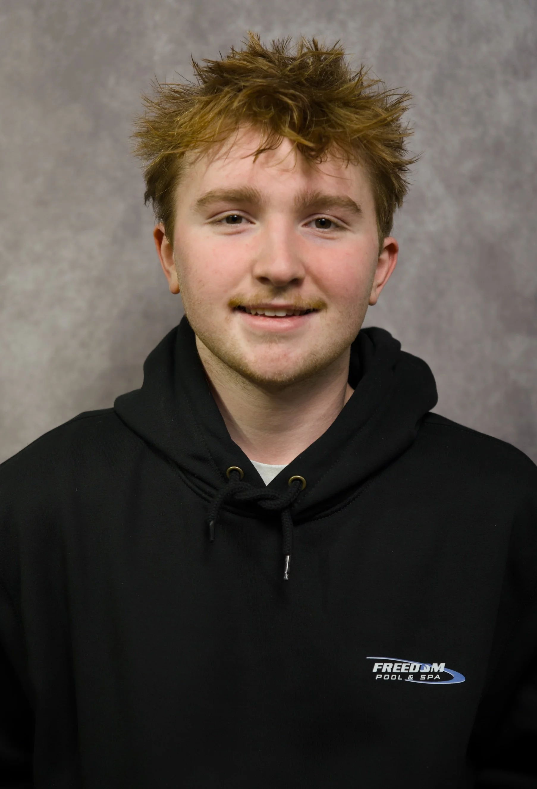 A young man with tousled light brown hair and a slight smile, wearing a black hoodie with a 'Freedom Pool & Spa' logo on it, standing against a gray textured background.