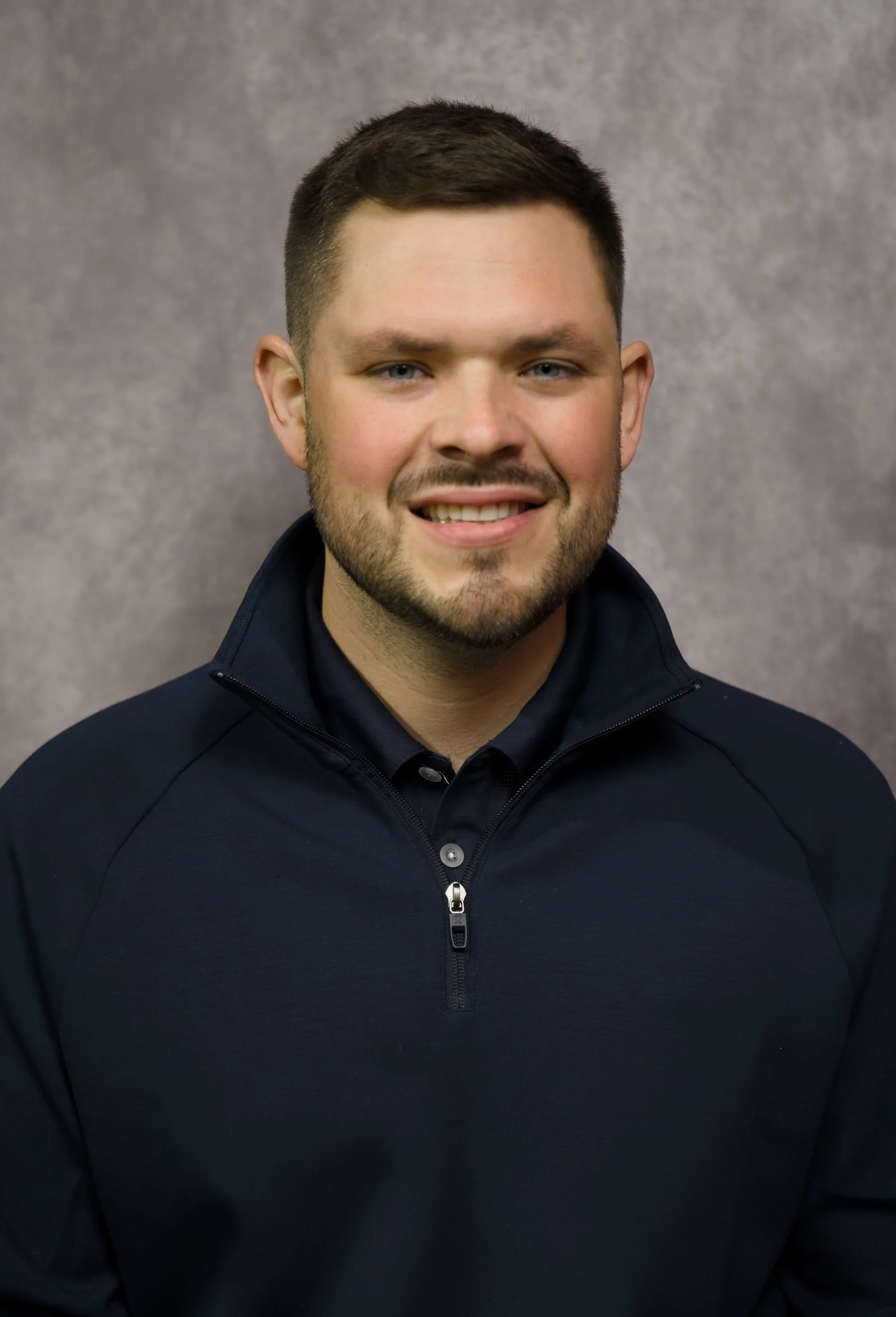 A young man with short dark hair and a beard smiling, wearing a dark zip-up jacket, against a gray background.