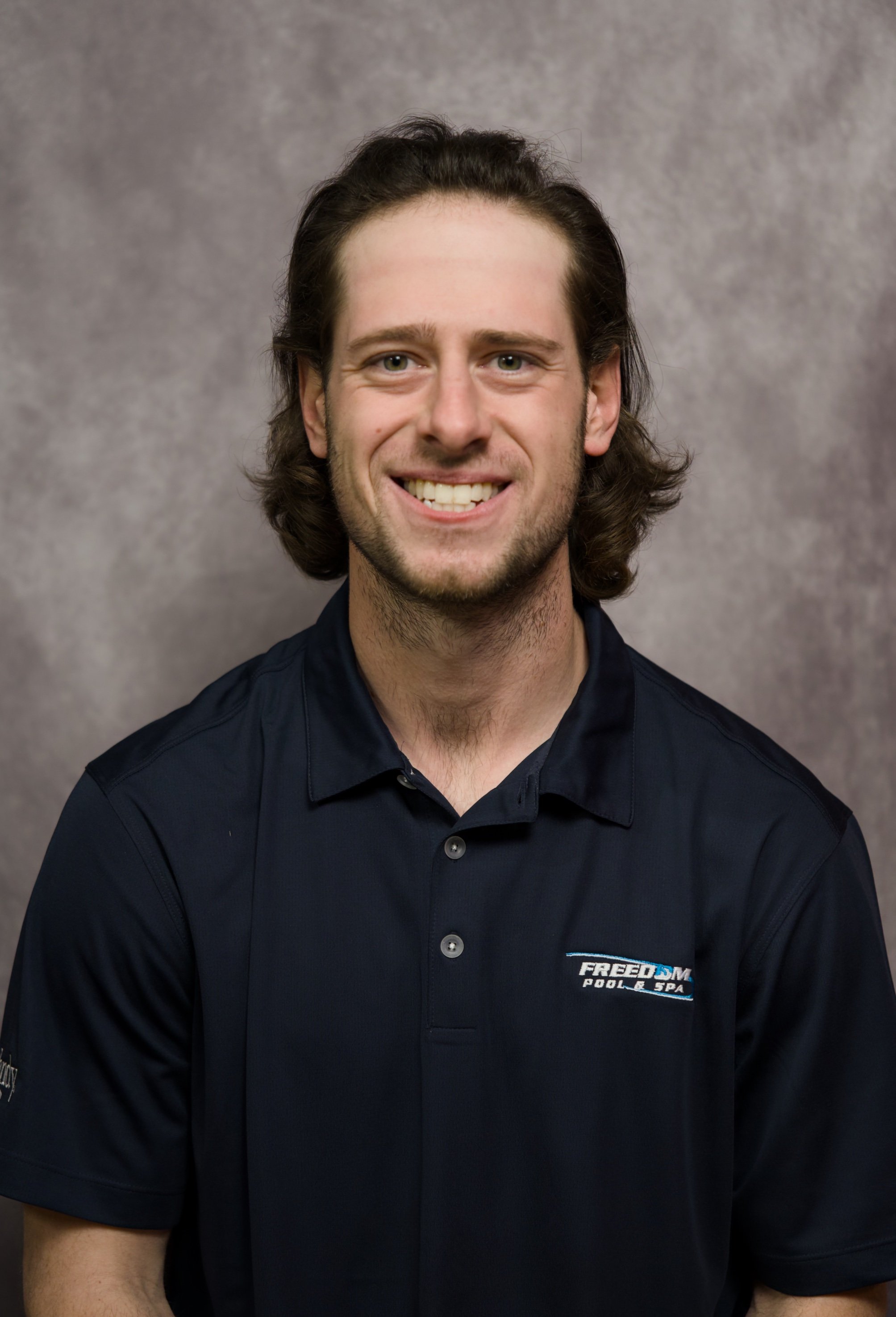 A smiling man with long brown hair, wearing a dark blue polo shirt with a logo that reads "FREEDOM POOL & SPA" on the left chest, standing against a gray background.